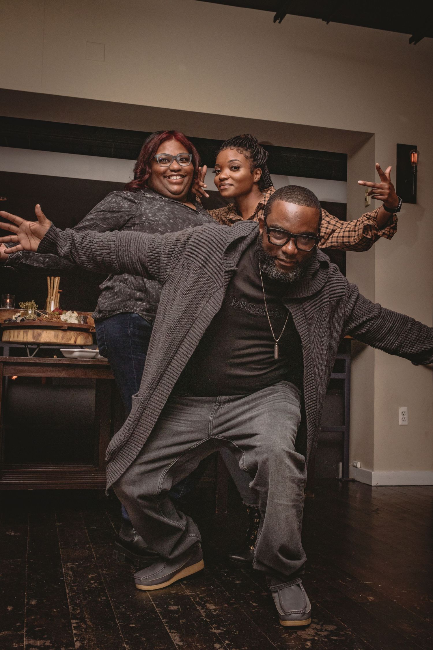 Three people wearing cozy sweaters strike playful poses while having fun on a wooden floor in a casual indoor setting.