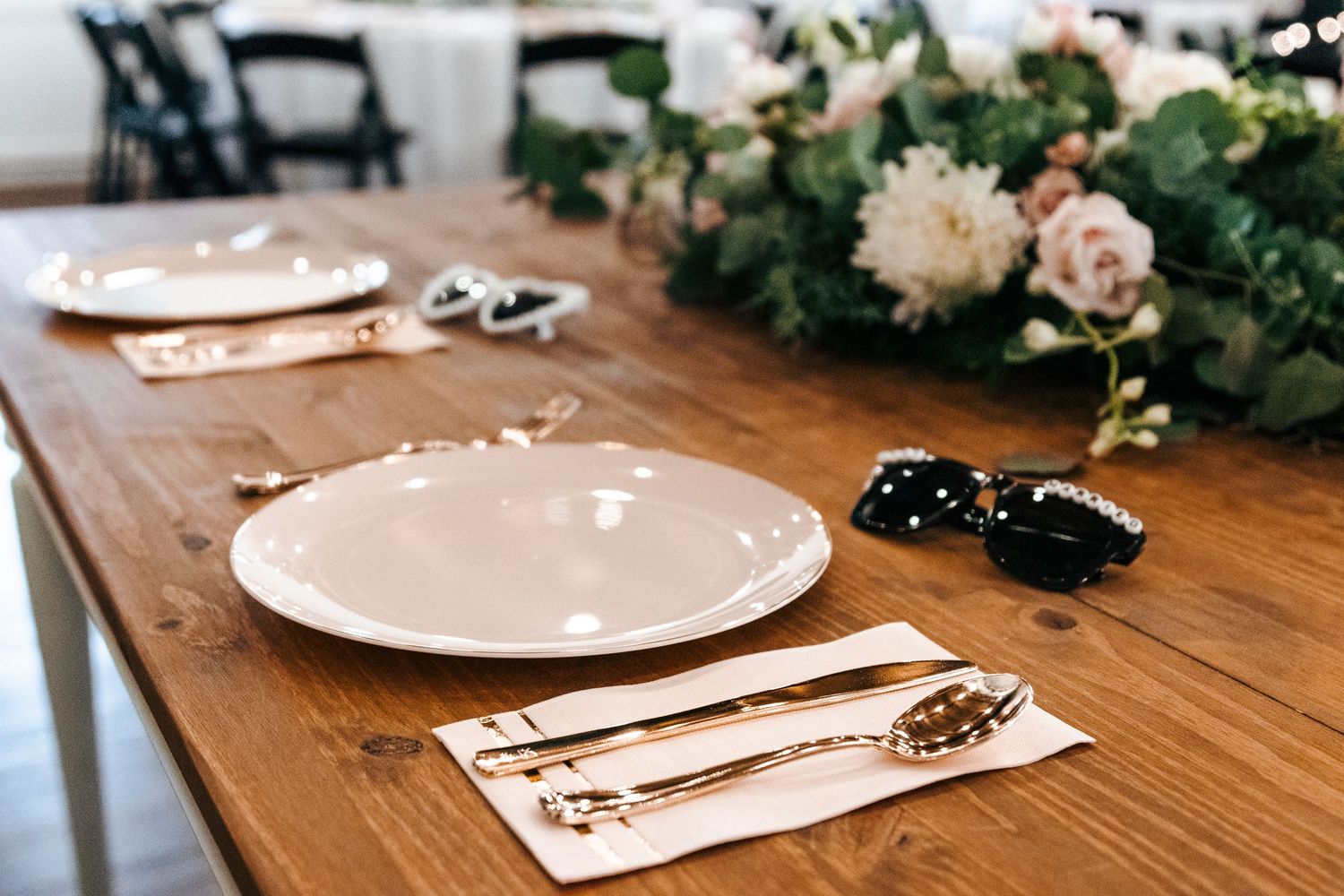 Elegant wooden table setting with white plates, gold cutlery, floral centerpiece, and sunglasses for a stylish event.