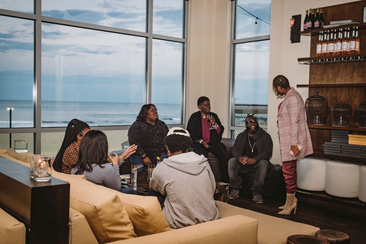 Groups of people sit at tables near large windows in a modern cafe overlooking an ocean view.