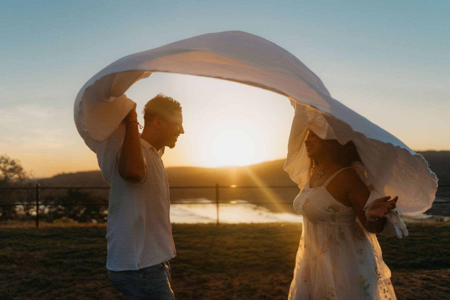 A couple holds up a flowing white veil against the golden sunset in a romantic outdoor photoshoot.