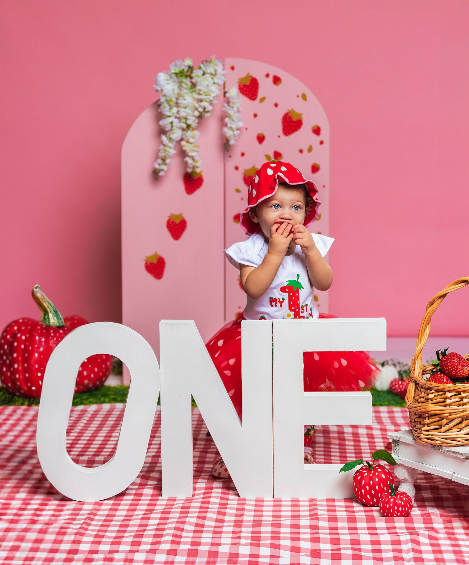 First birthday photo setup with pink backdrop, large 'ONE' letters and strawberry themed decorations.