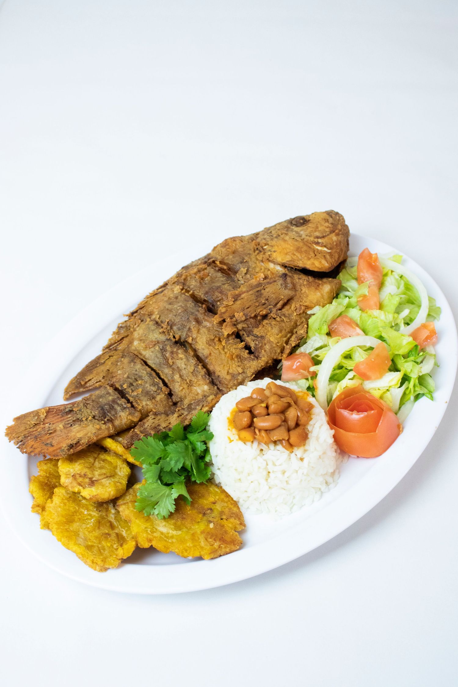 Fried fish platter with rice, plantains and fresh salad on a white plate against light background.