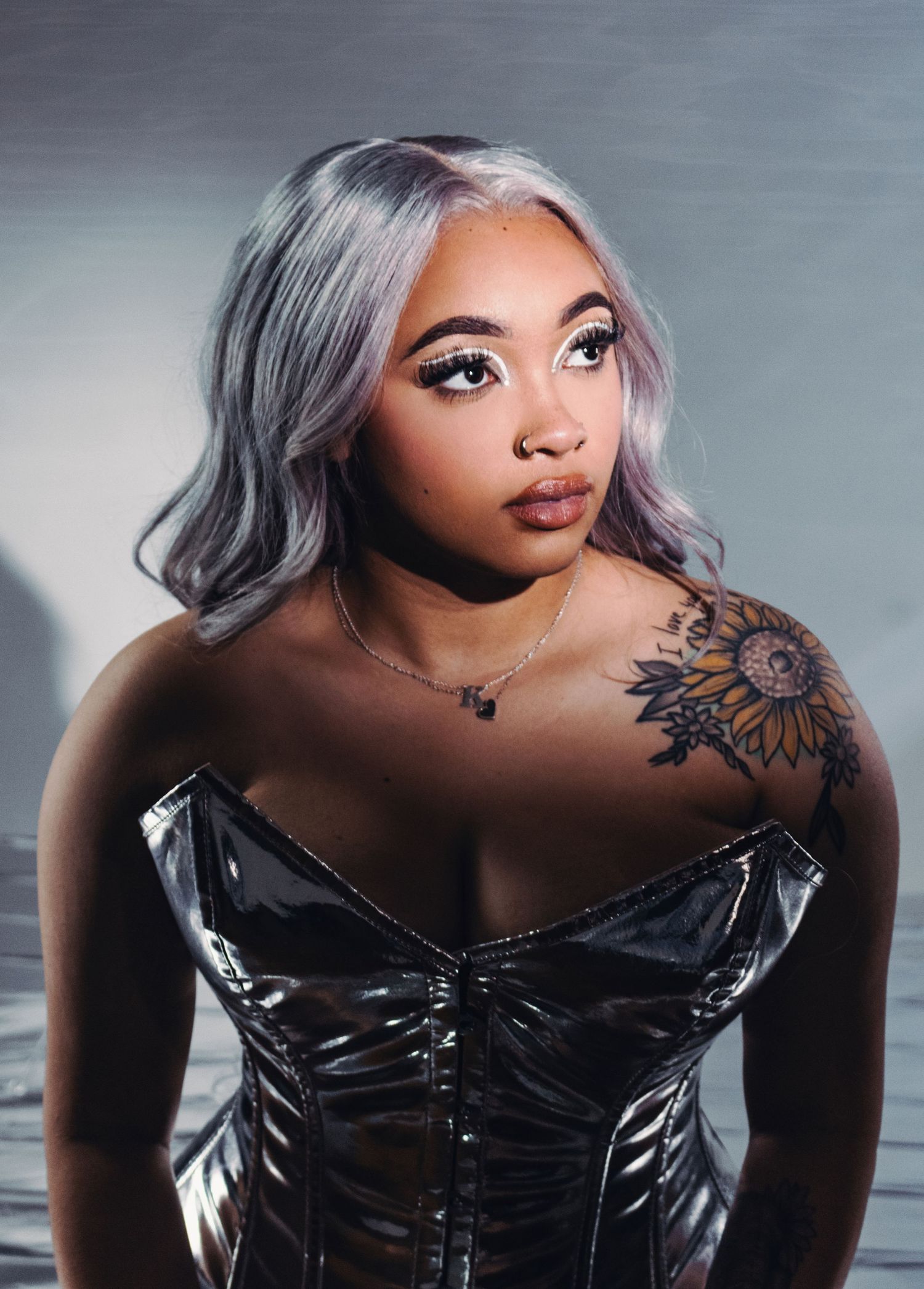 Portrait showing silver hair and black corset with shoulder tattoo against gray background.