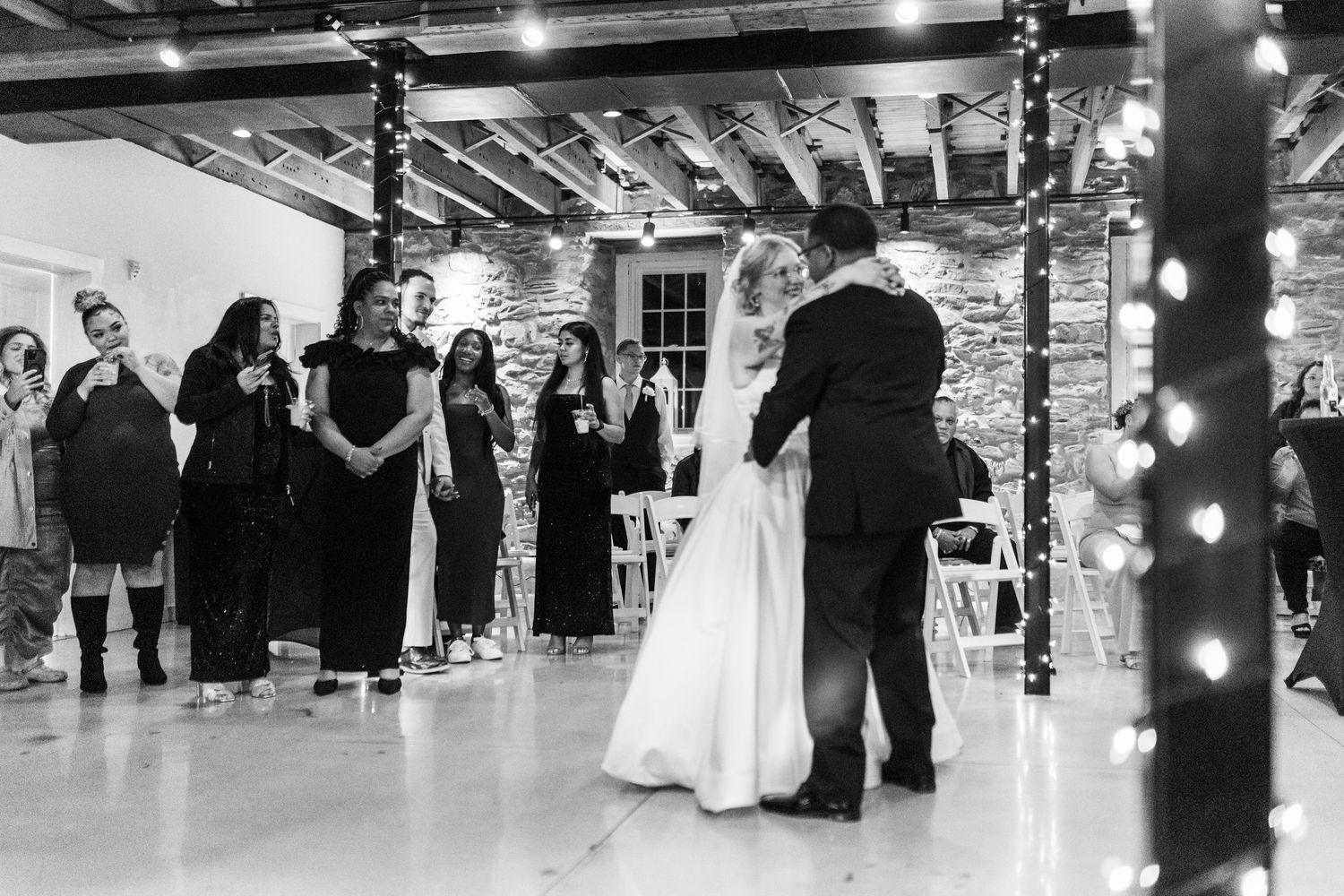 Bride and groom share first dance in rustic stone venue with fairy lights while guests watch in black and white photo.