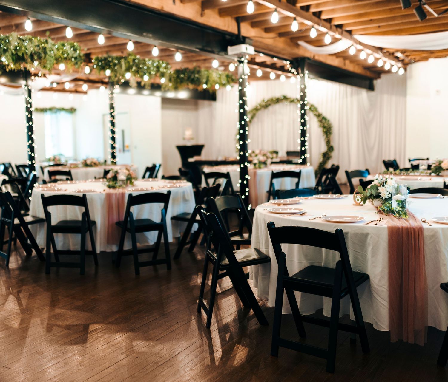 Elegant wedding reception hall with black folding chairs, round tables, string lights, and greenery garland decor.