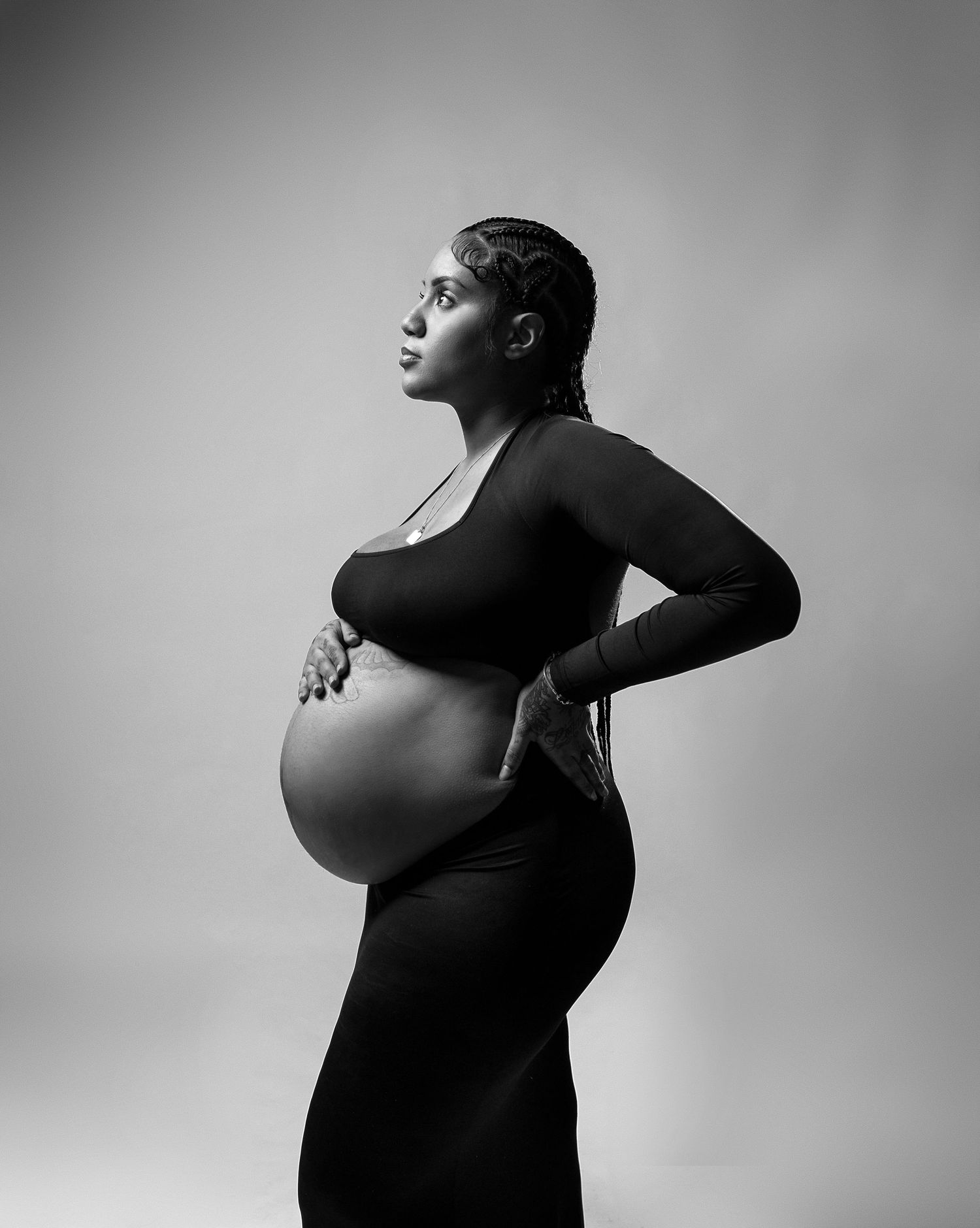 Side profile of pregnant woman in black dress against grey background in artistic black and white portrait.