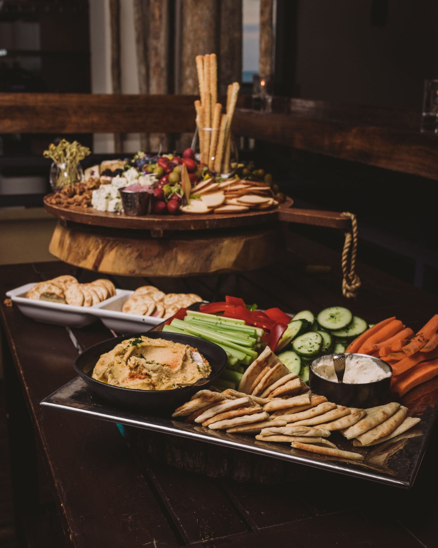 Variety of appetizers and crudités artfully arranged on dark wooden charcuterie boards with bread sticks and dips.