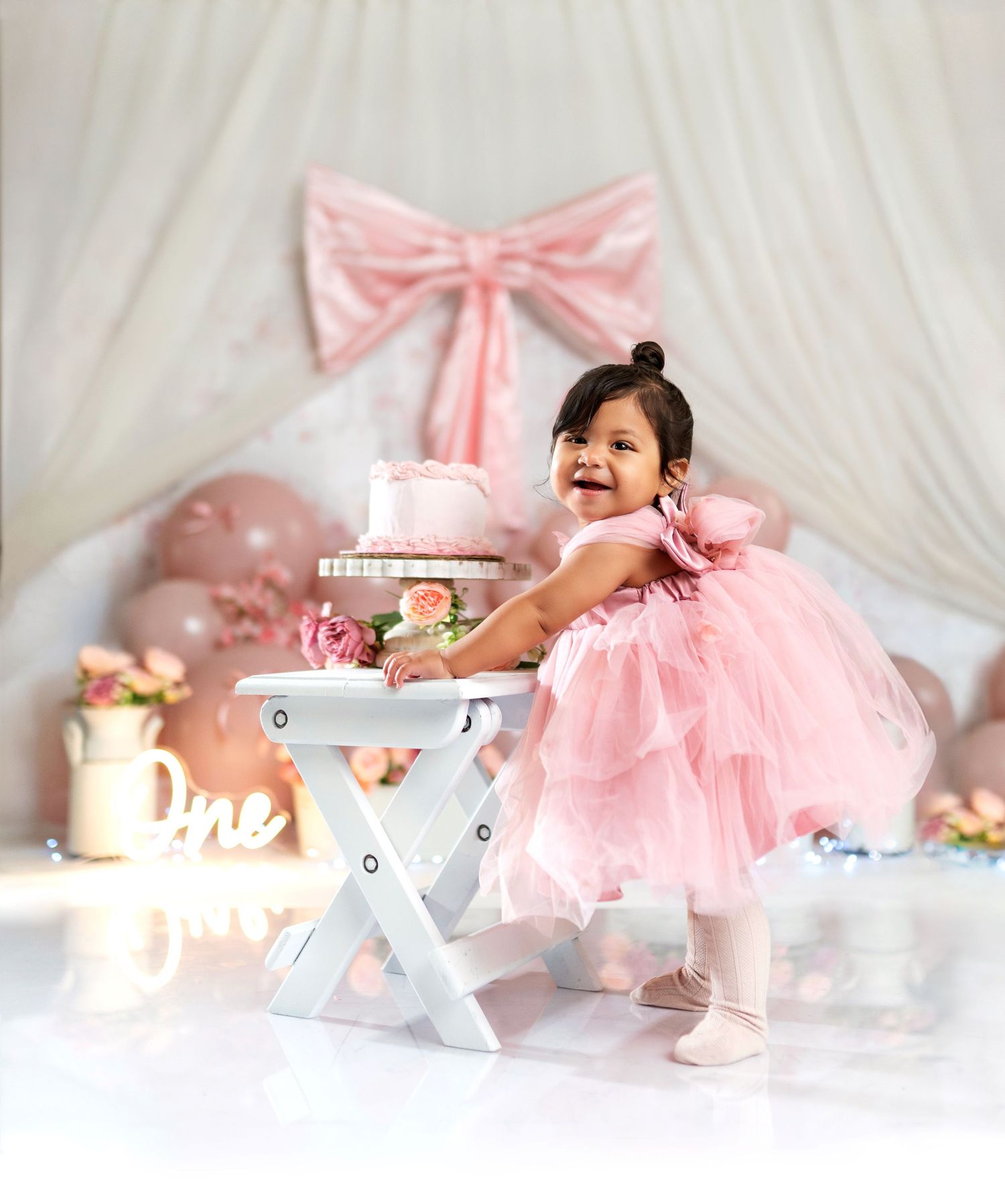 Baby girl in pink tutu dress smiles at first birthday cake smash setup with balloons and bow backdrop.