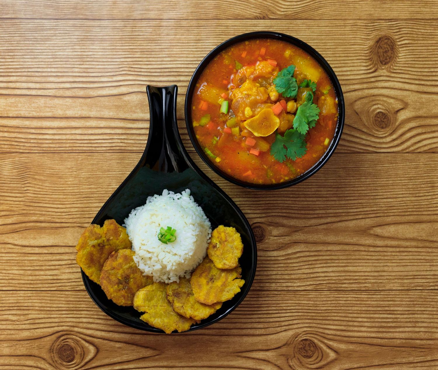Curry dish with rice and fried plantains served in black bowls on wooden table.
