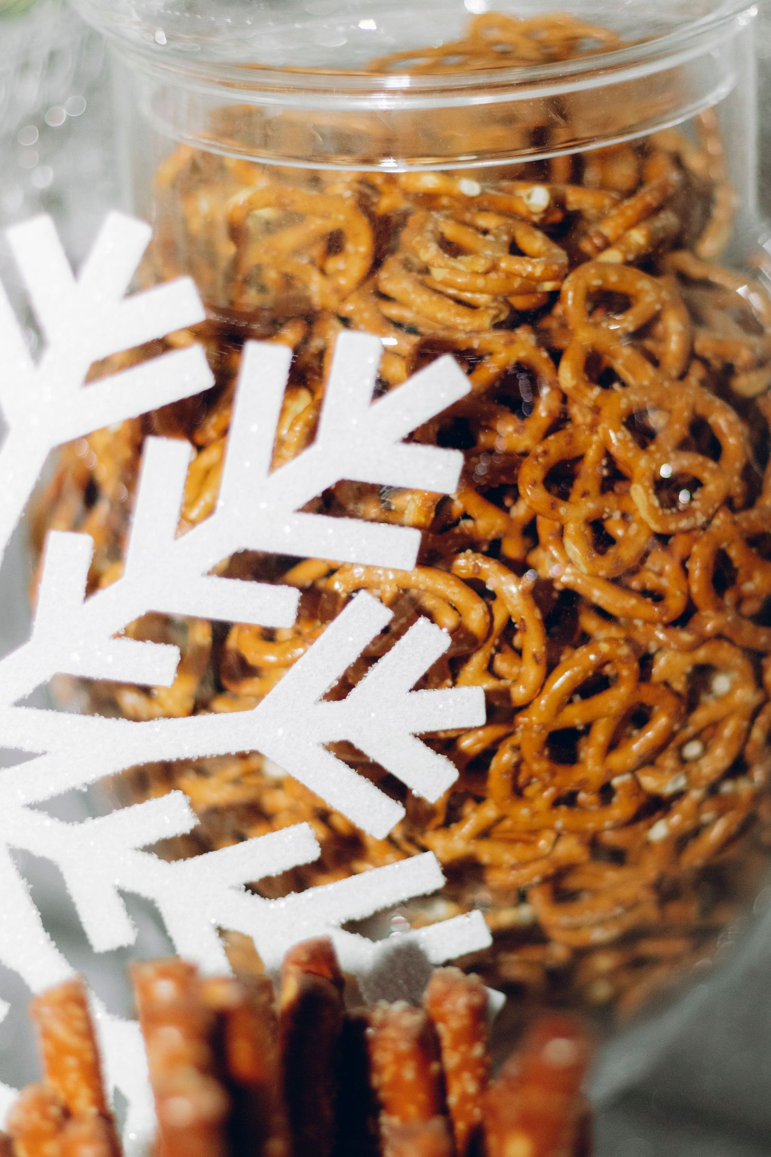 Glass jar filled with crunchy brown pretzel snacks decorated with white snowflake shapes for winter holiday season.