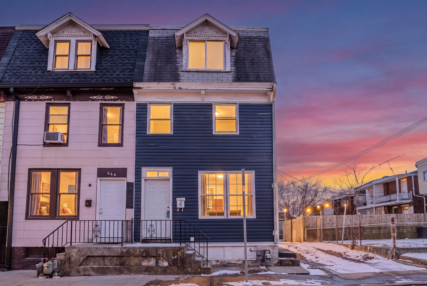 Navy blue townhouse exterior glowing warmly at dusk with snow on ground and vibrant pink and purple sunset sky.