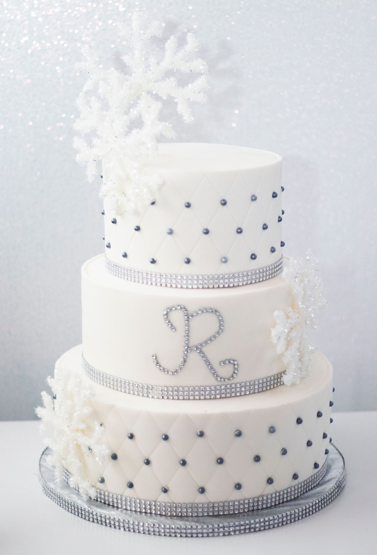 Elegant white three-tier wedding cake decorated with silver crystals, snowflakes, and a rhinestone monogram letter R.