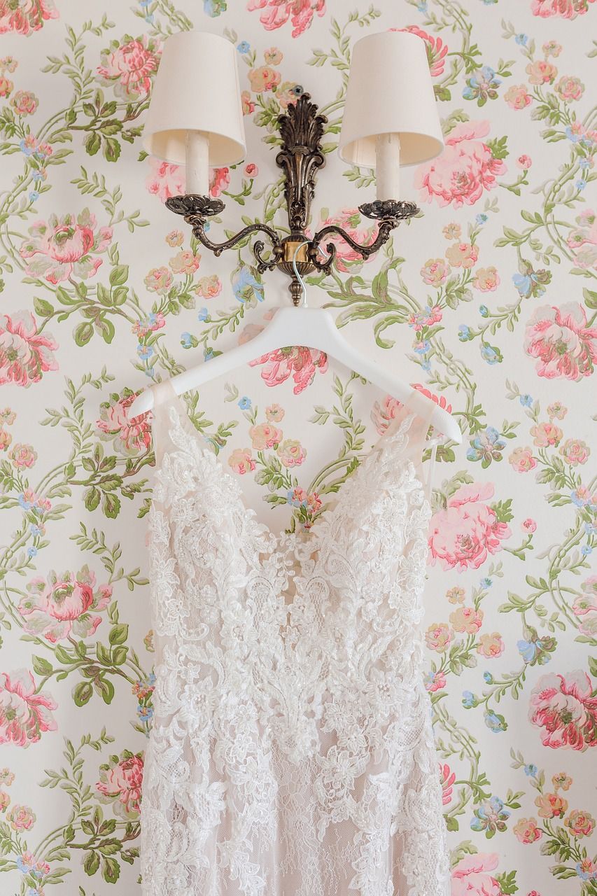 Three elegant white lace wedding dresses hang against floral pink and green vintage wallpaper in a bridal shop.