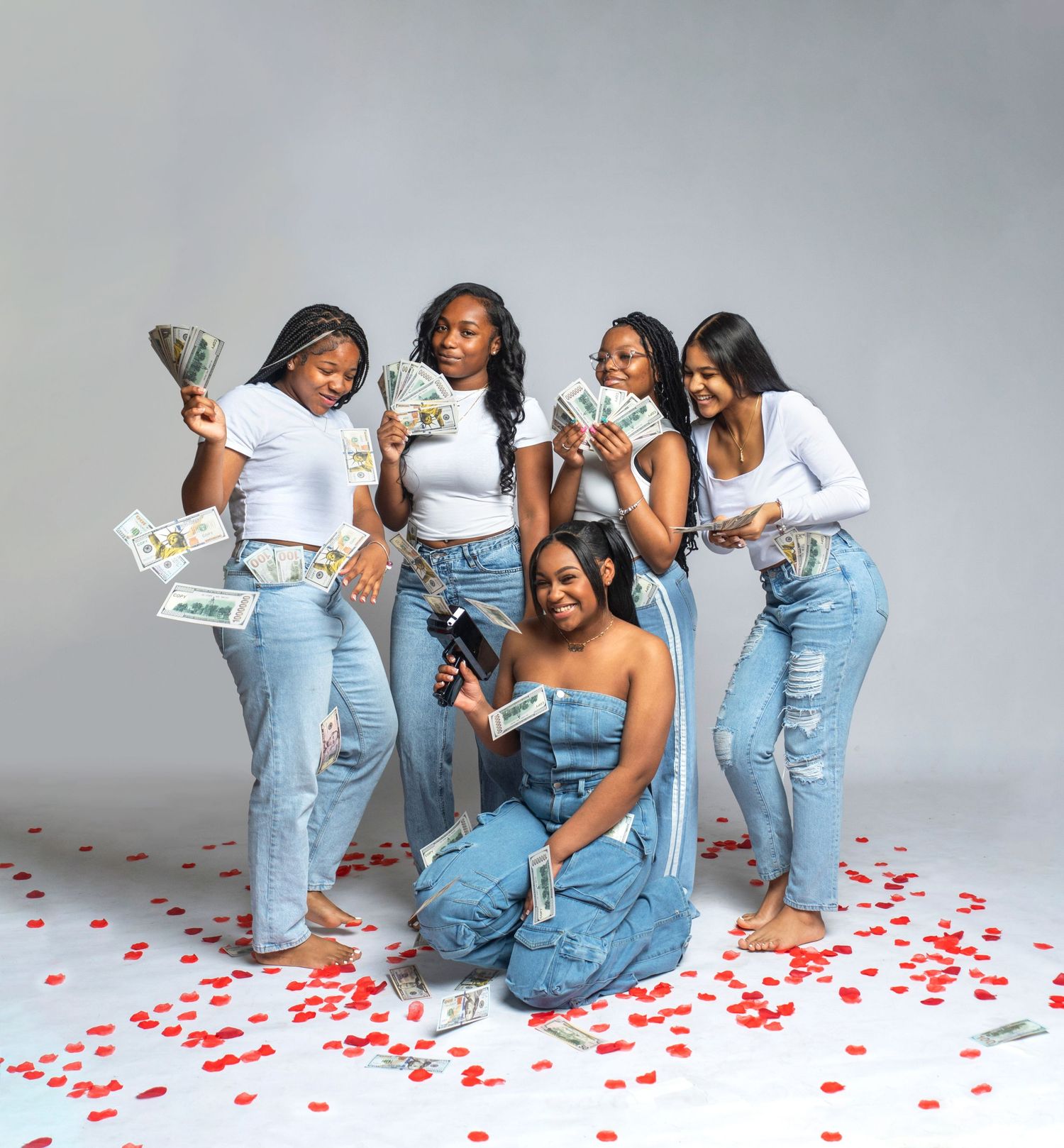 Playful fashion photoshoot with models in denim jeans posing with money against a gray backdrop with red petals.