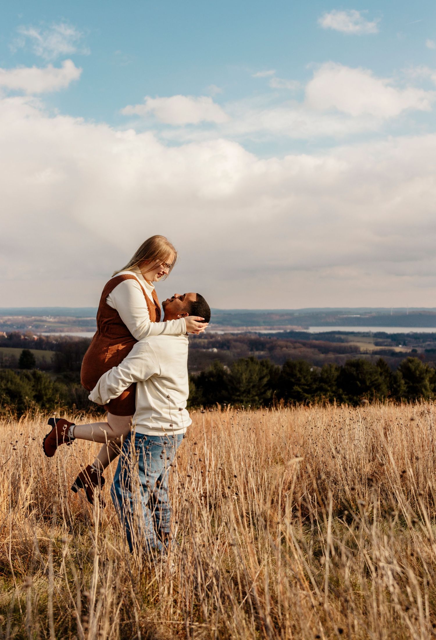 A playful couple enjoys a romantic moment in a golden wheat field on a sunny day with blue skies and white clouds.
