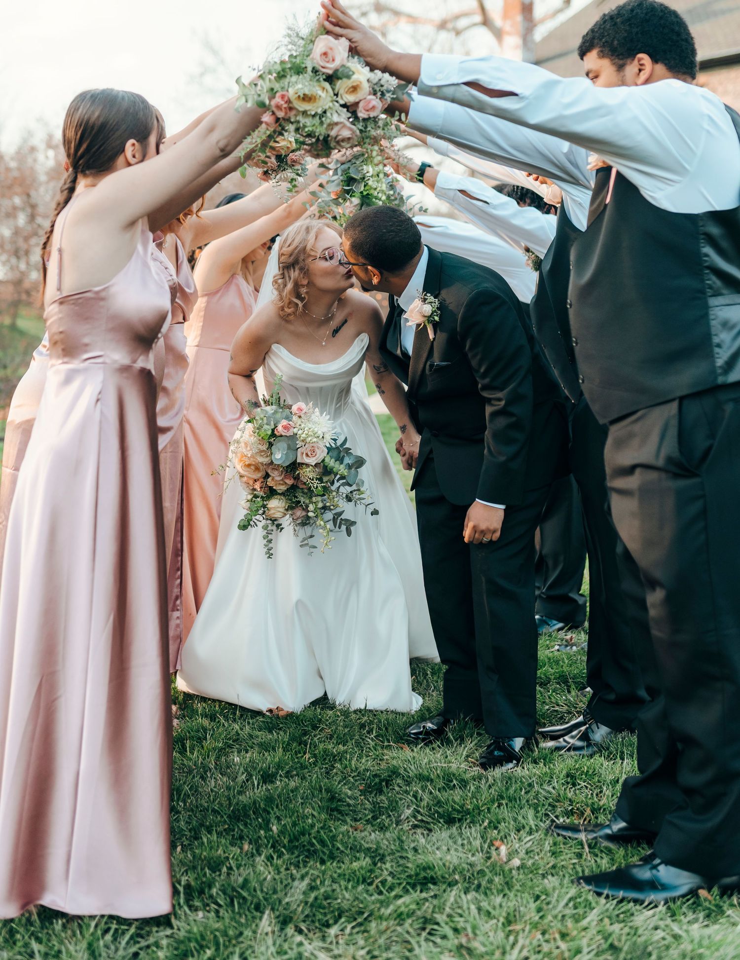 Bride and groom kiss under an arch of flowers and ribbons held by wedding party on a grassy lawn.