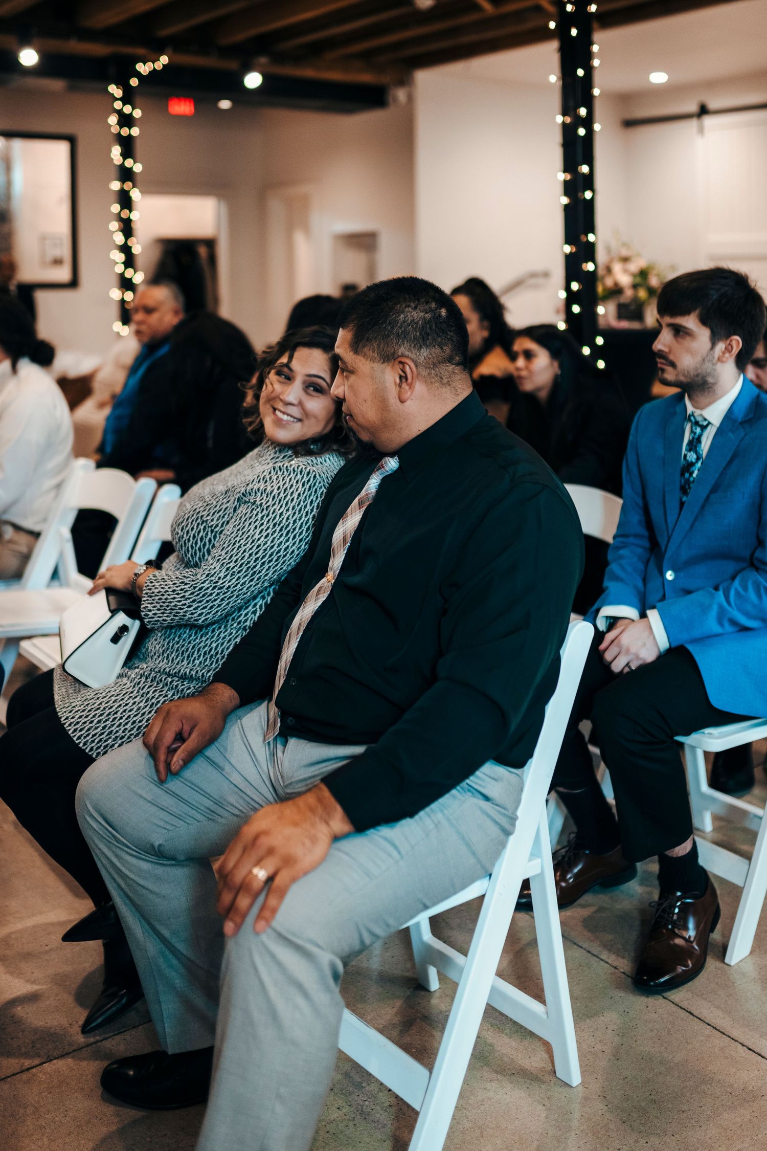 Wedding guests share a tender moment seated in white chairs during an indoor ceremony with warm string lights.