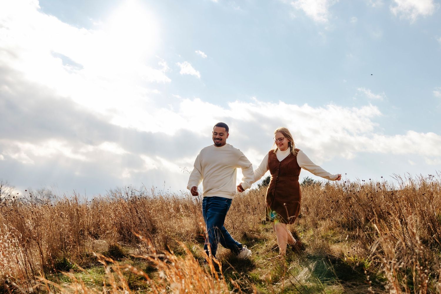 A couple runs joyfully through a golden wheat field under cloudy skies at sunset.