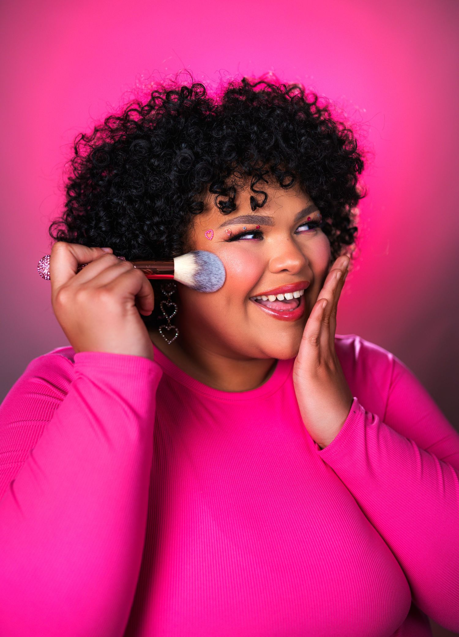A makeup artist wearing a bright pink sweater applies cosmetics against a magenta background in sequential poses.