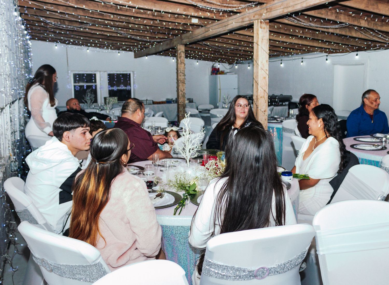 People sitting at a long white table at an outdoor dinner party under string lights and a wooden pergola.