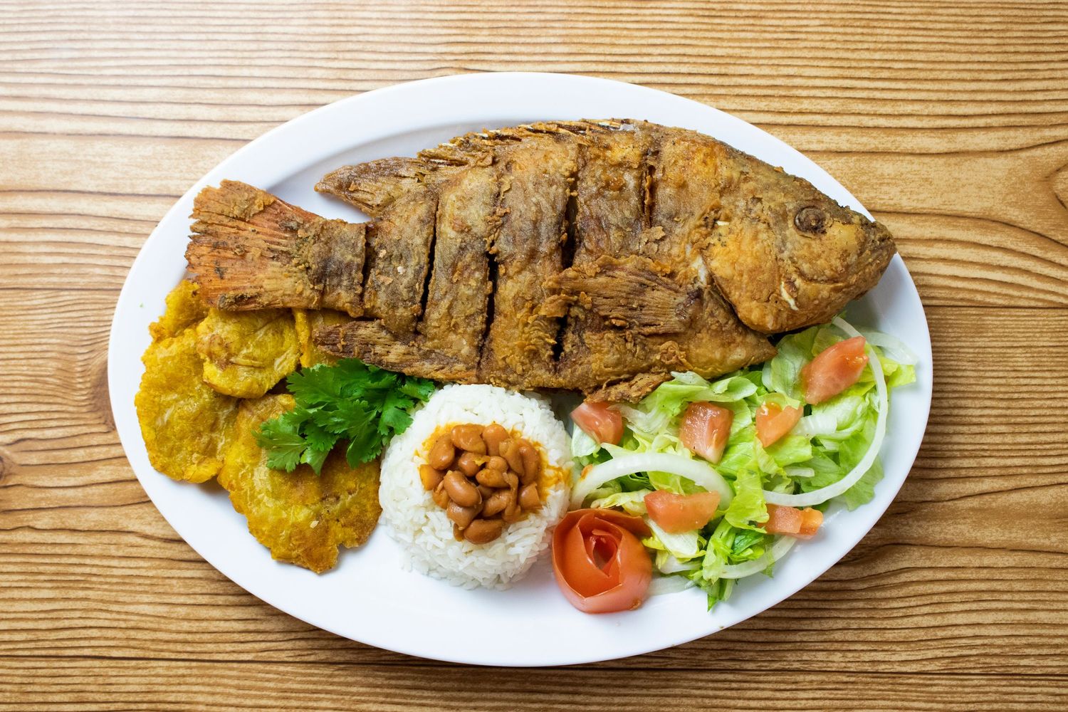 Grilled whole fish served with yellow plantains, white rice, fresh salad and garnish on a wooden table.