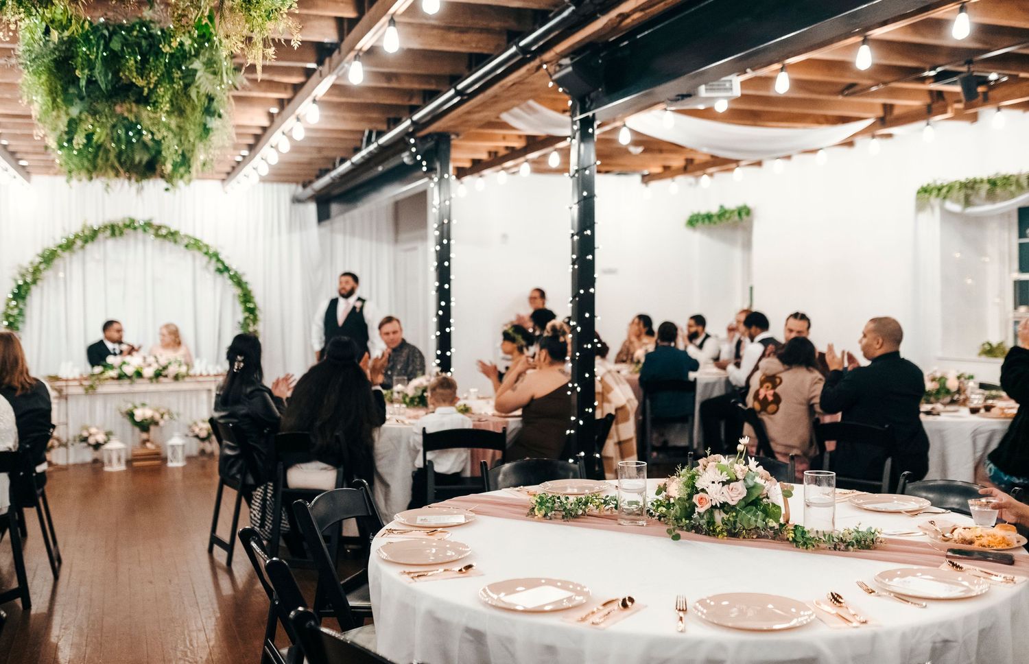 Elegant indoor wedding reception with round tables, floral centerpieces, and string lights on exposed ceiling.