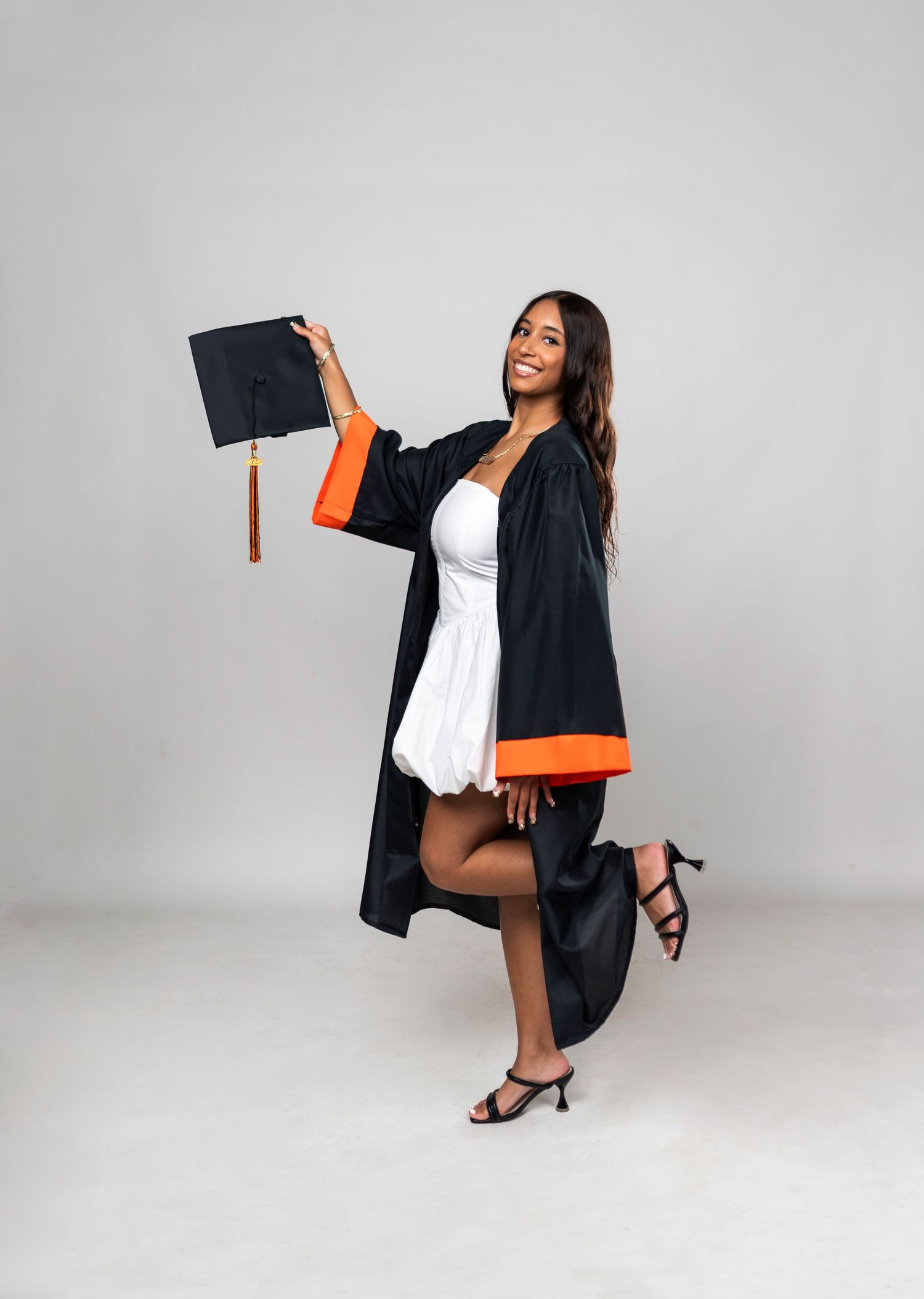 Graduate in white dress and black graduation gown celebrates while kicking up one leg.