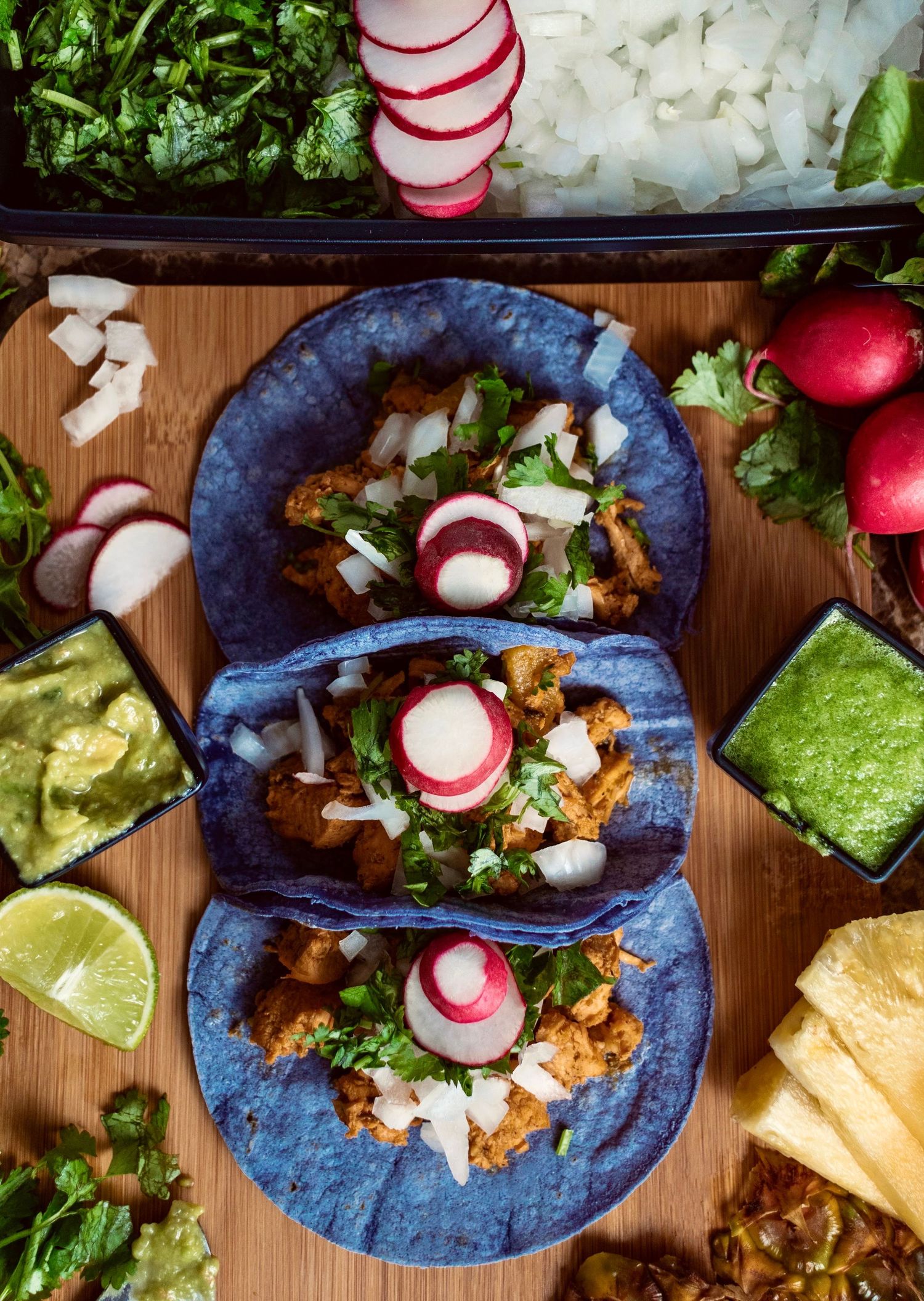 Blue corn tortilla tacos topped with radishes, cilantro and onions on wooden serving board.