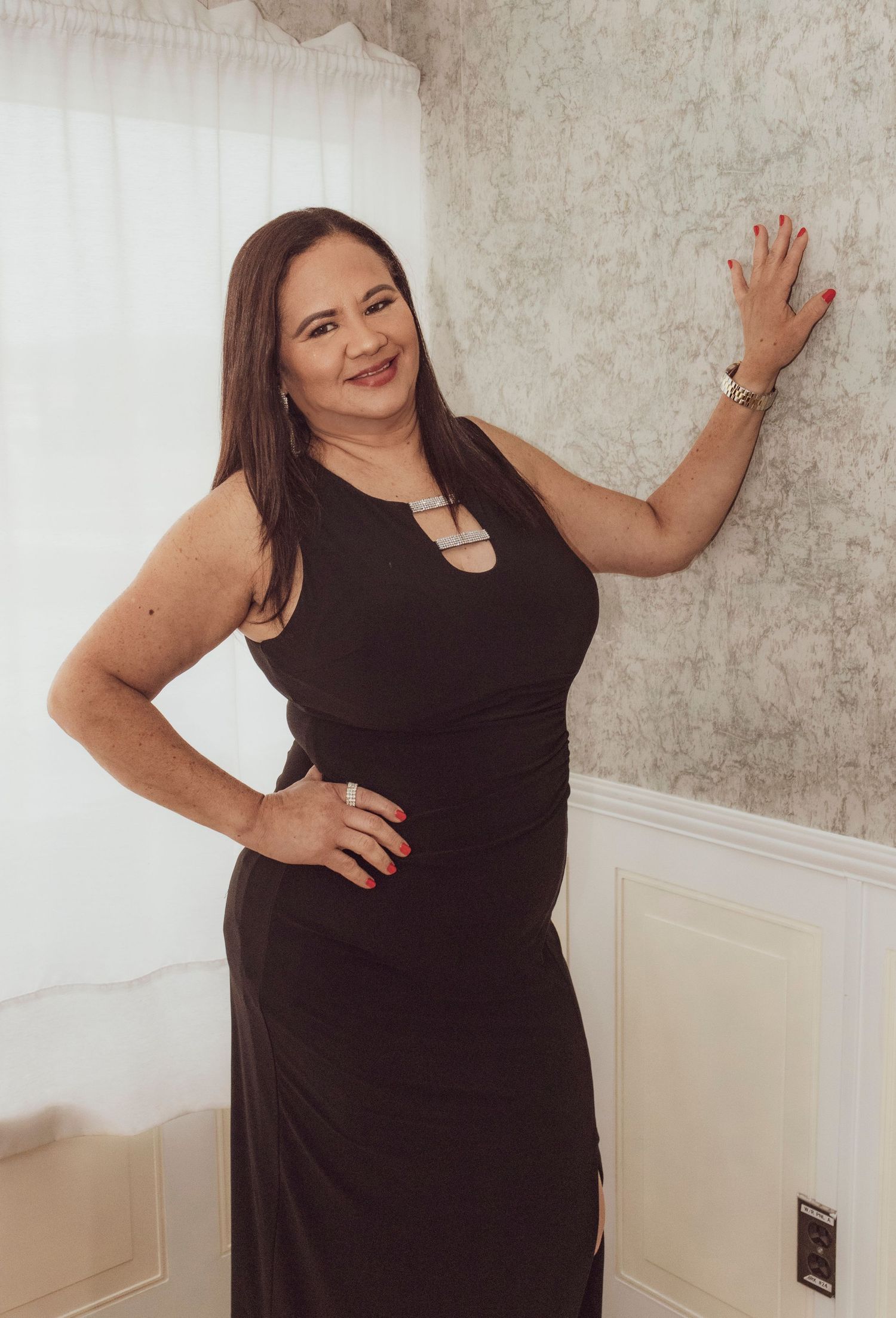 Woman in a black formal dress poses confidently against a light colored wall.