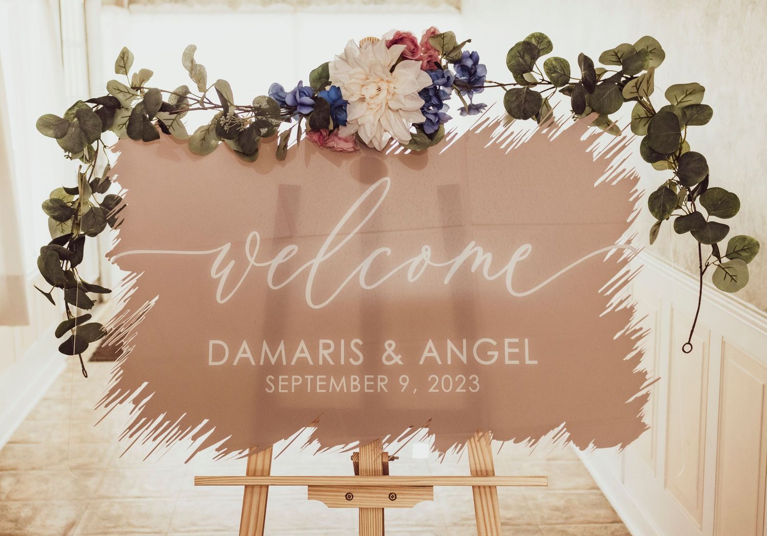 Rustic wedding welcome sign decorated with greenery and flowers displays the names Damaris & Angel.