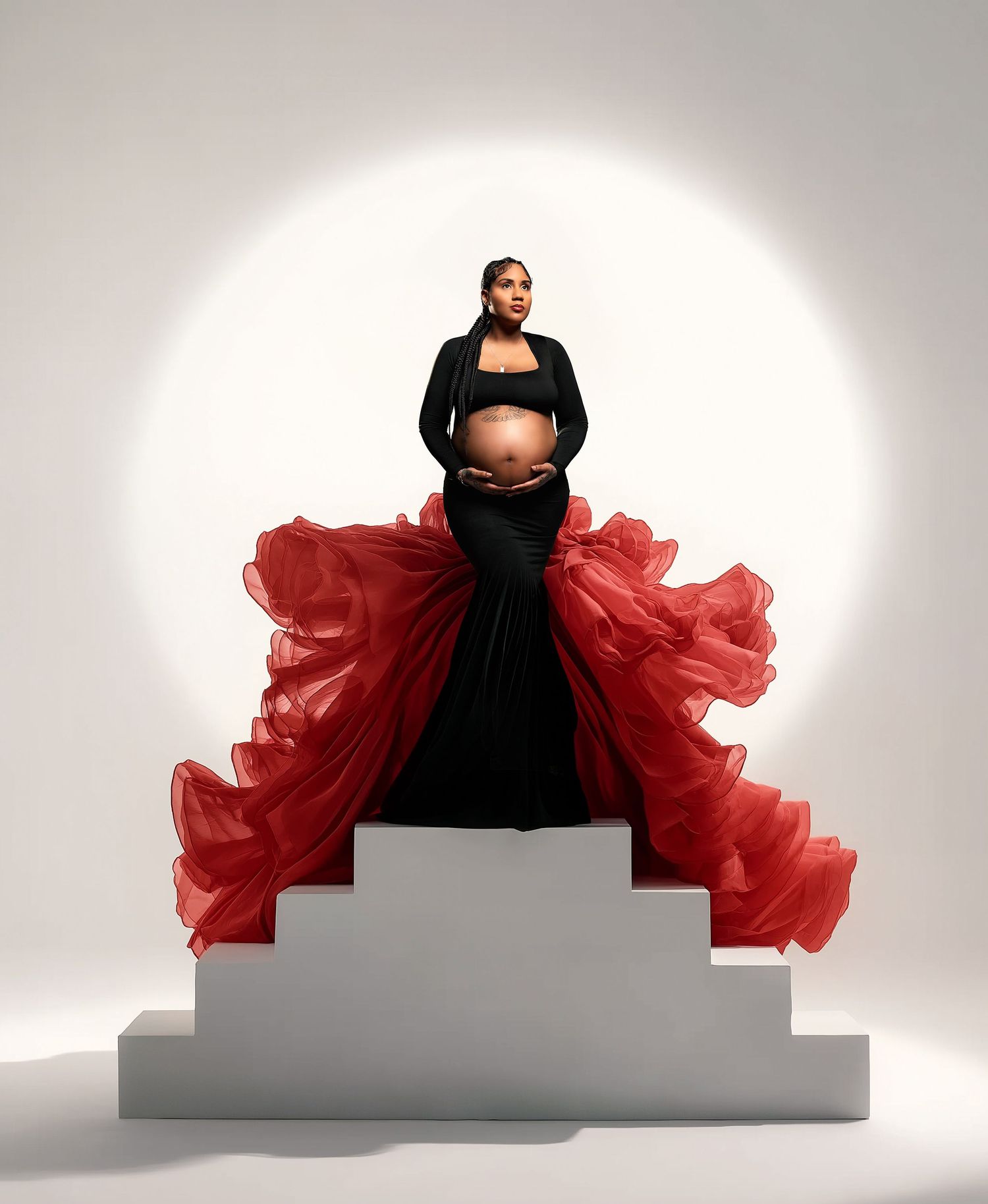 A fashion model in a dramatic black and red tulle gown poses on a white platform against a light backdrop.