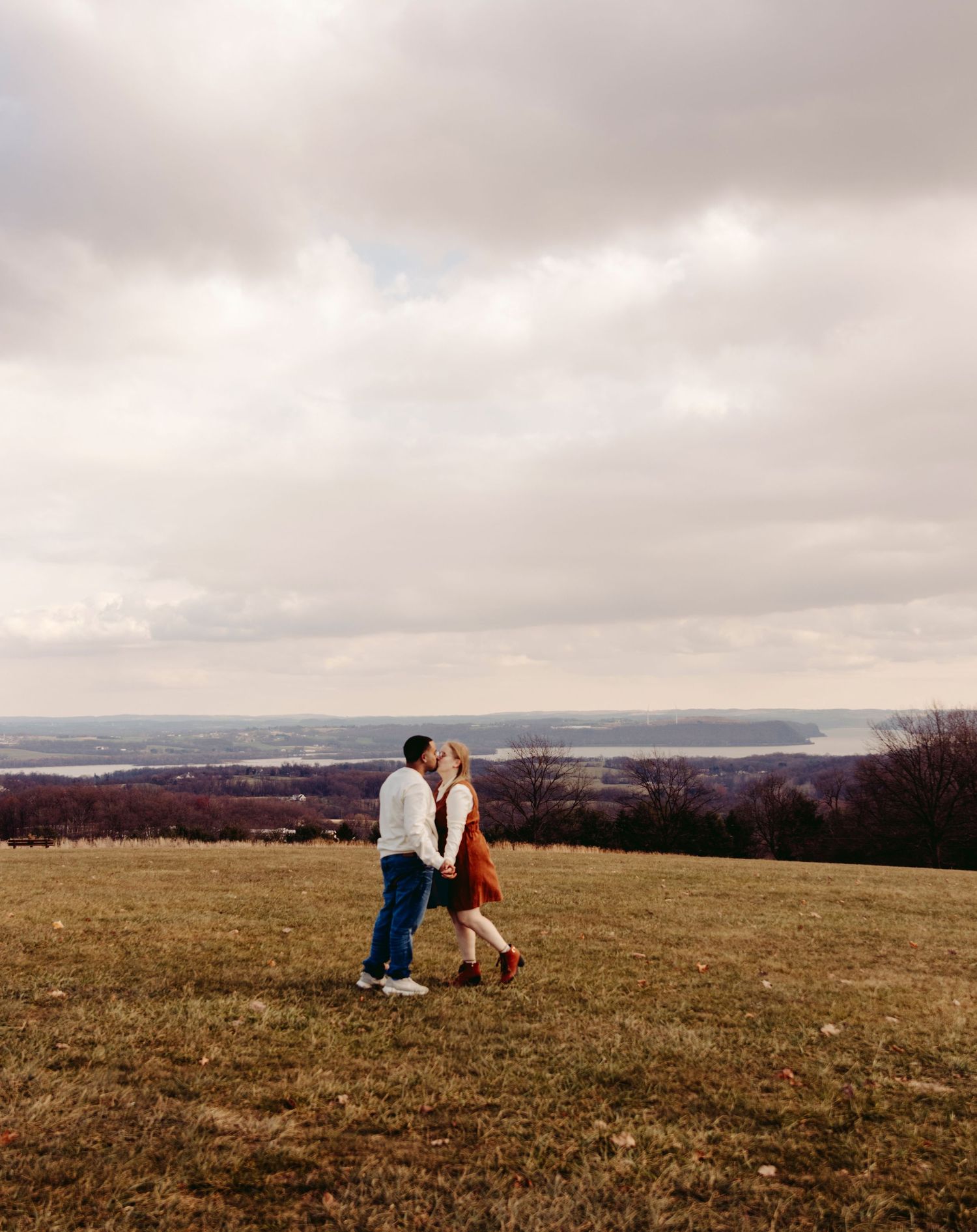 Two figures sit close together on a rolling hillside under a dramatic cloudy sky at sunset.
