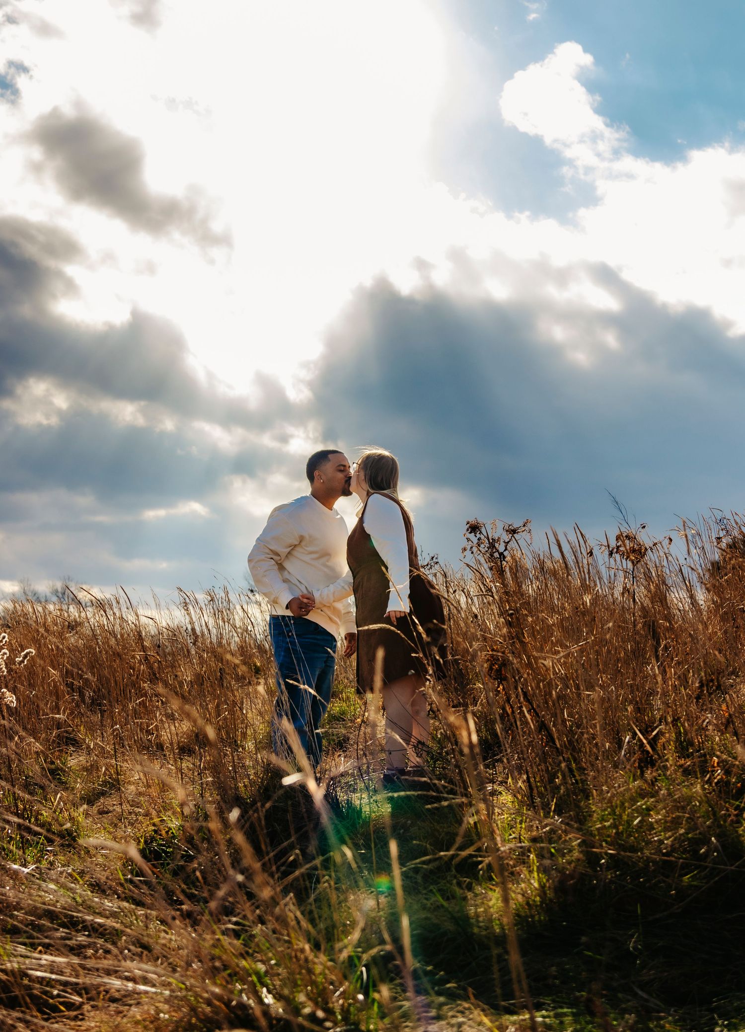 A couple embraces on a hilltop with golden grass and dramatic clouds in the background at sunset.