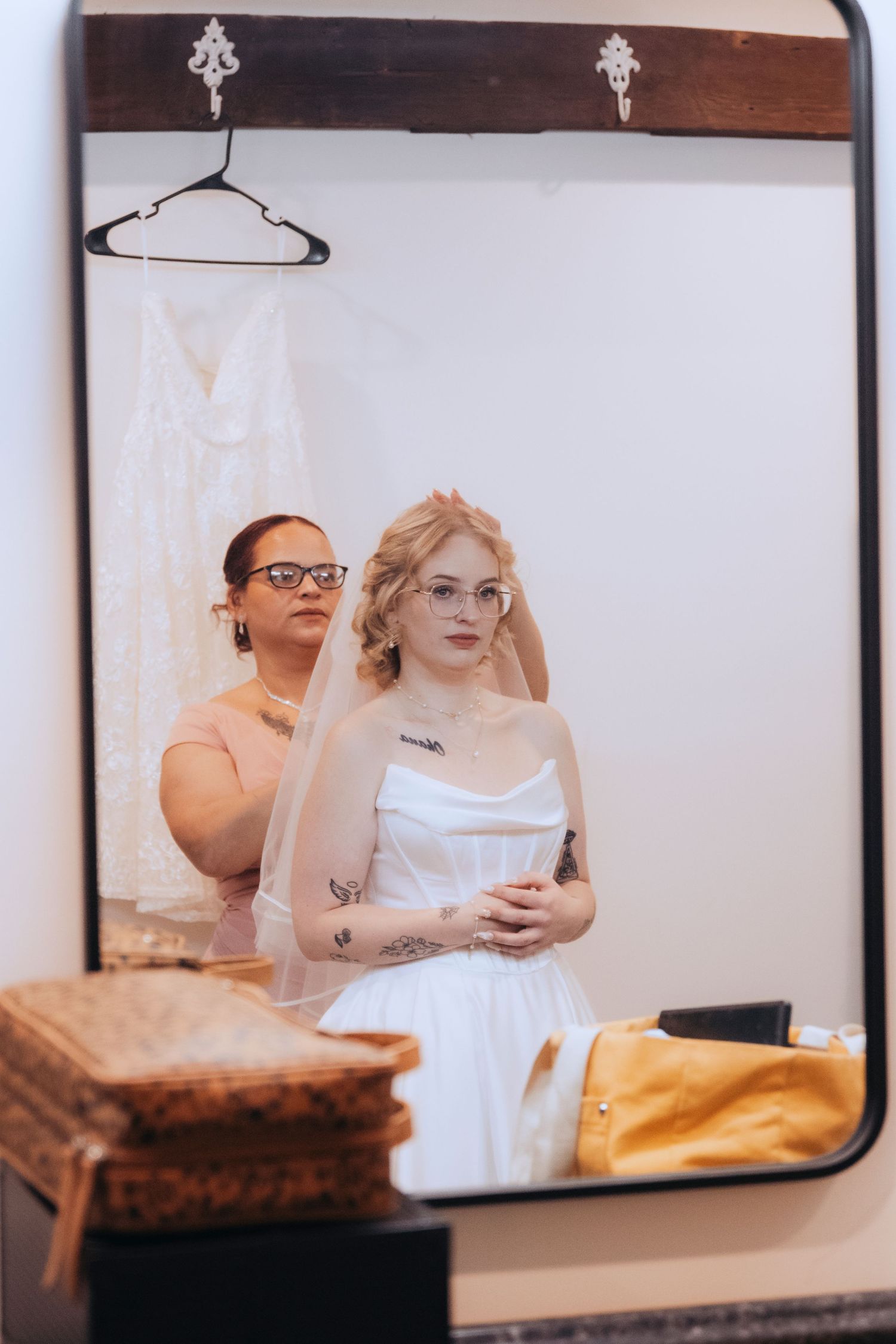 Bride in white gown and veil gets ready as bridesmaid adjusts her hair, reflected in a vintage mirror.