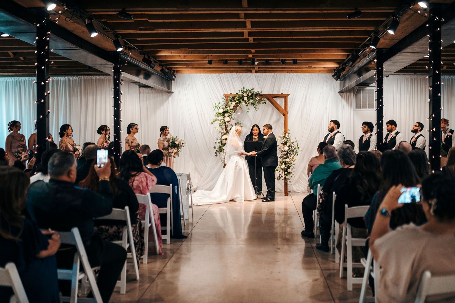 Wedding ceremony in rustic venue with wooden arch, white draping, and seated guests.
