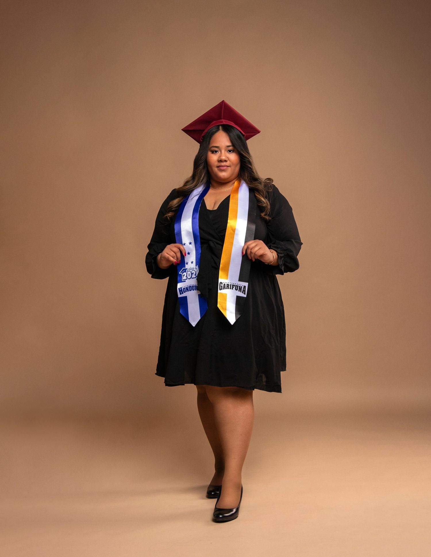 Graduate in black cap and gown with stoles poses against brown backdrop for professional photo.