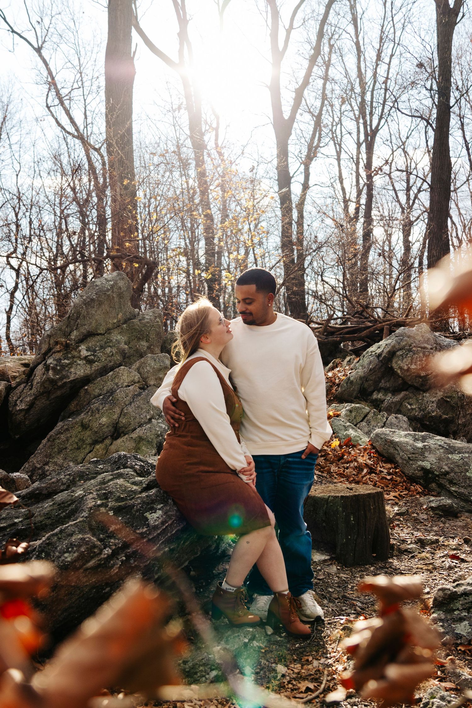 A couple sits together on large rocks in a sunlit autumn forest setting with fallen leaves surrounding them.