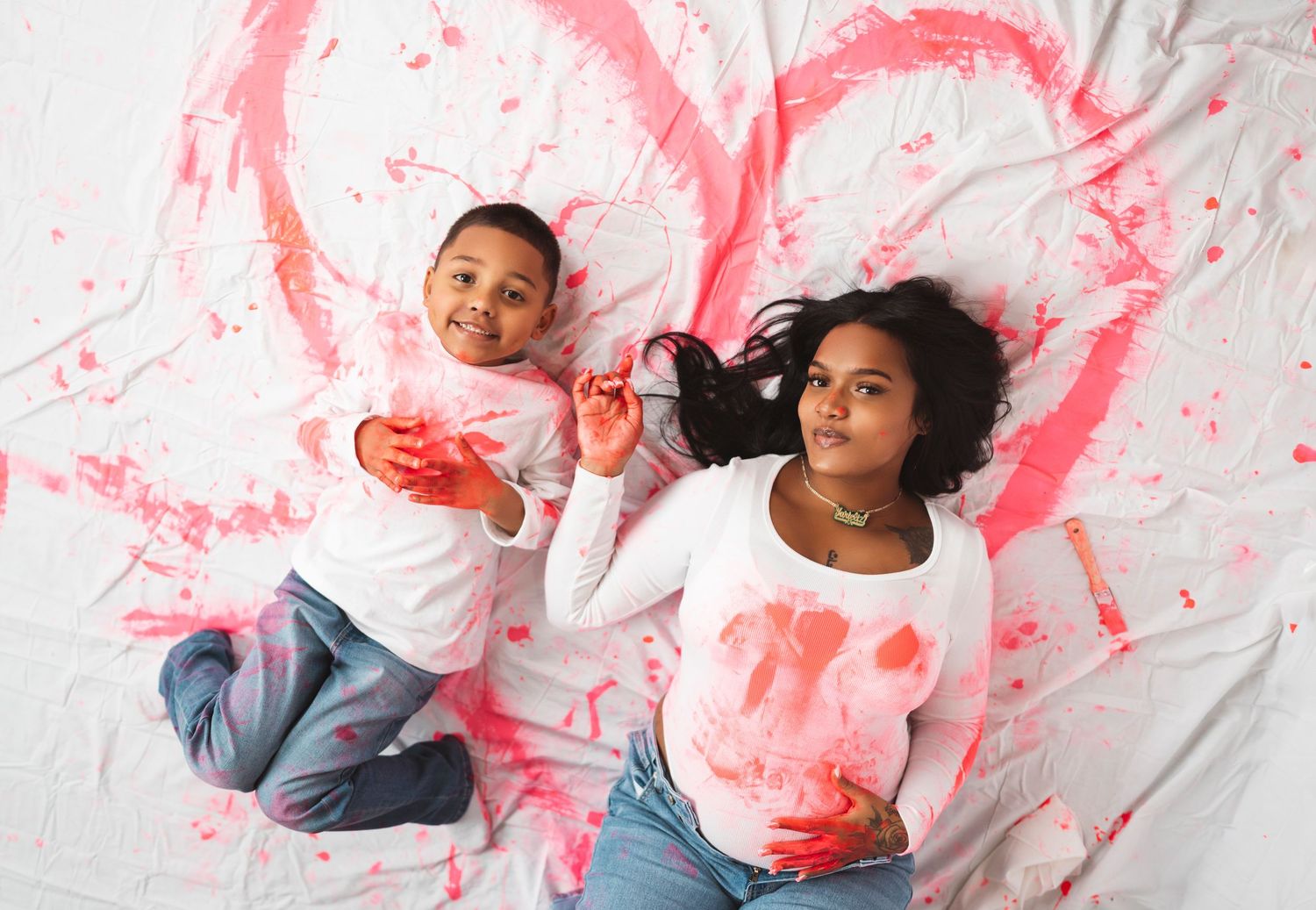 Two children lying on white sheet with pink painted heart, holding paintbrushes, smiling at camera.