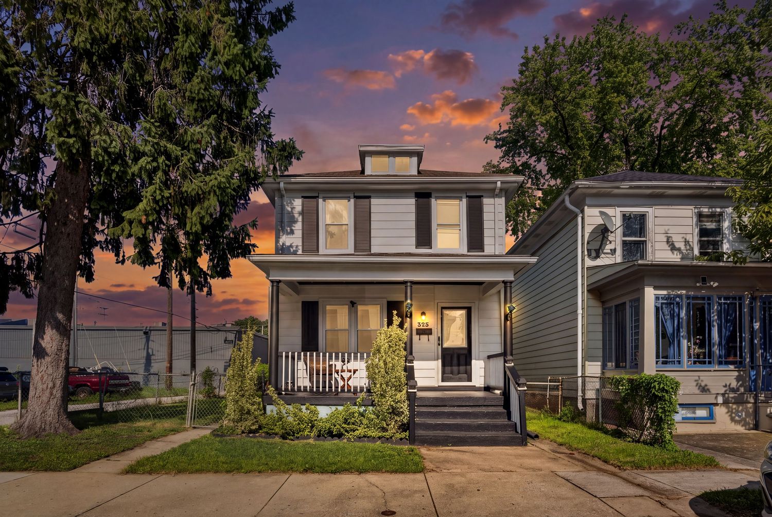 Gray two-story home with covered porch, lush trees, and a stunning sunset sky in a residential neighborhood.