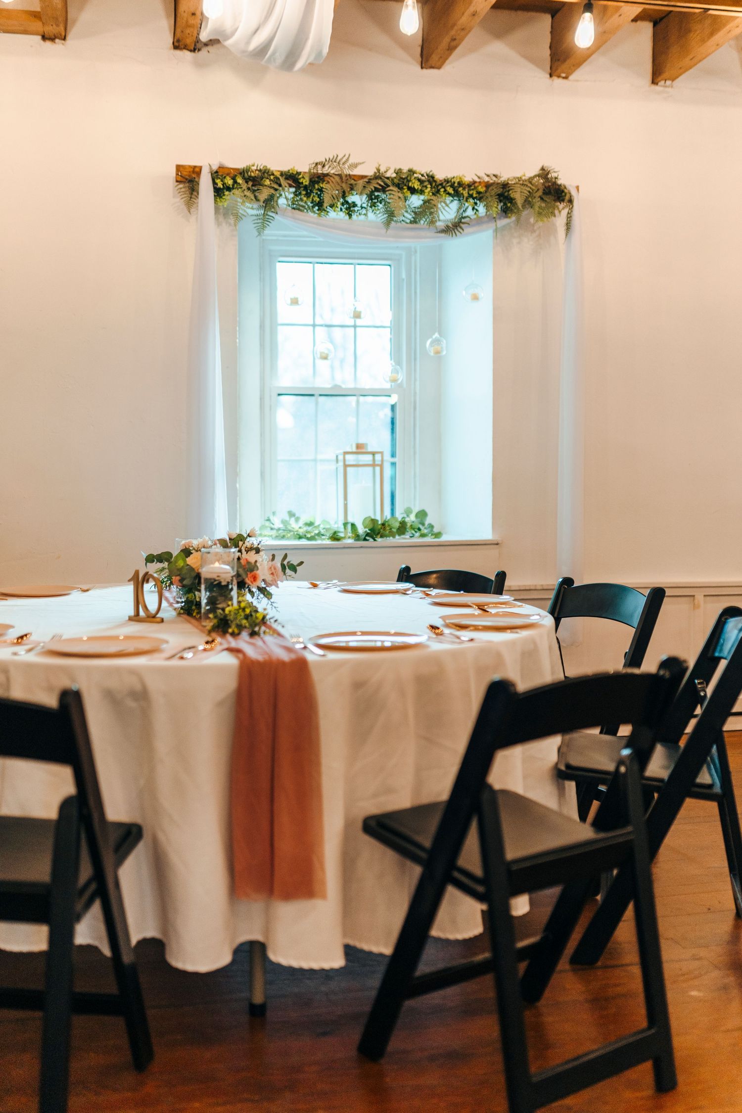 Elegant wedding reception table with white linen, floral centerpiece, black folding chairs, and greenery garland above window.