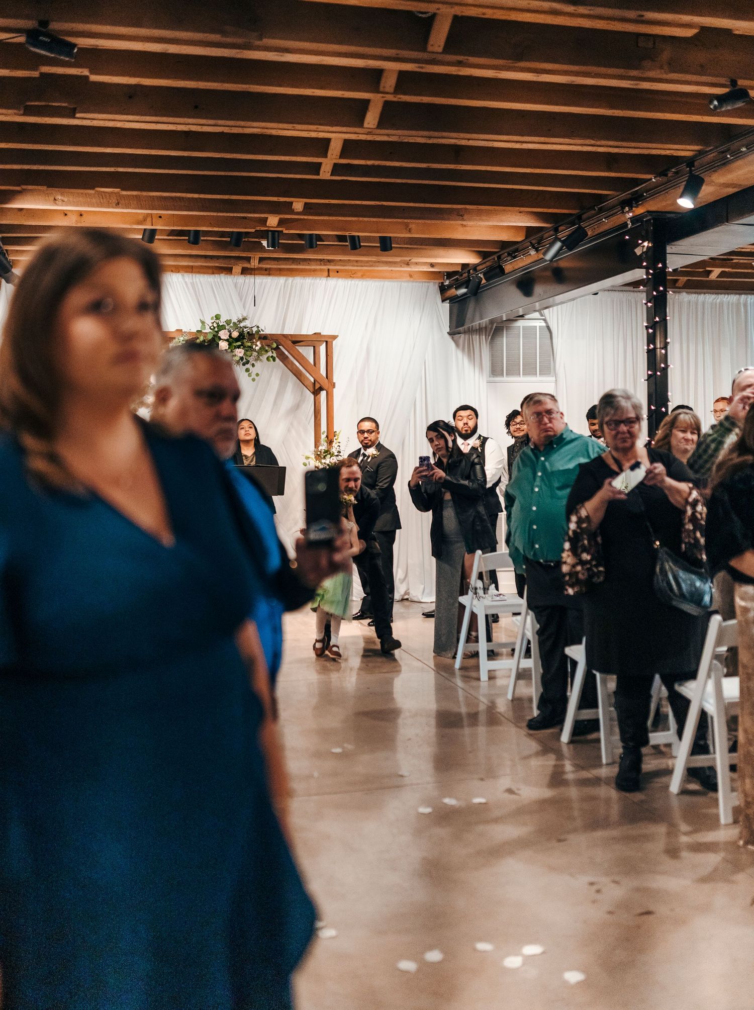 Guests stand in a rustic indoor wedding venue with wood beam ceilings, white chairs, and a floral arch at the altar.