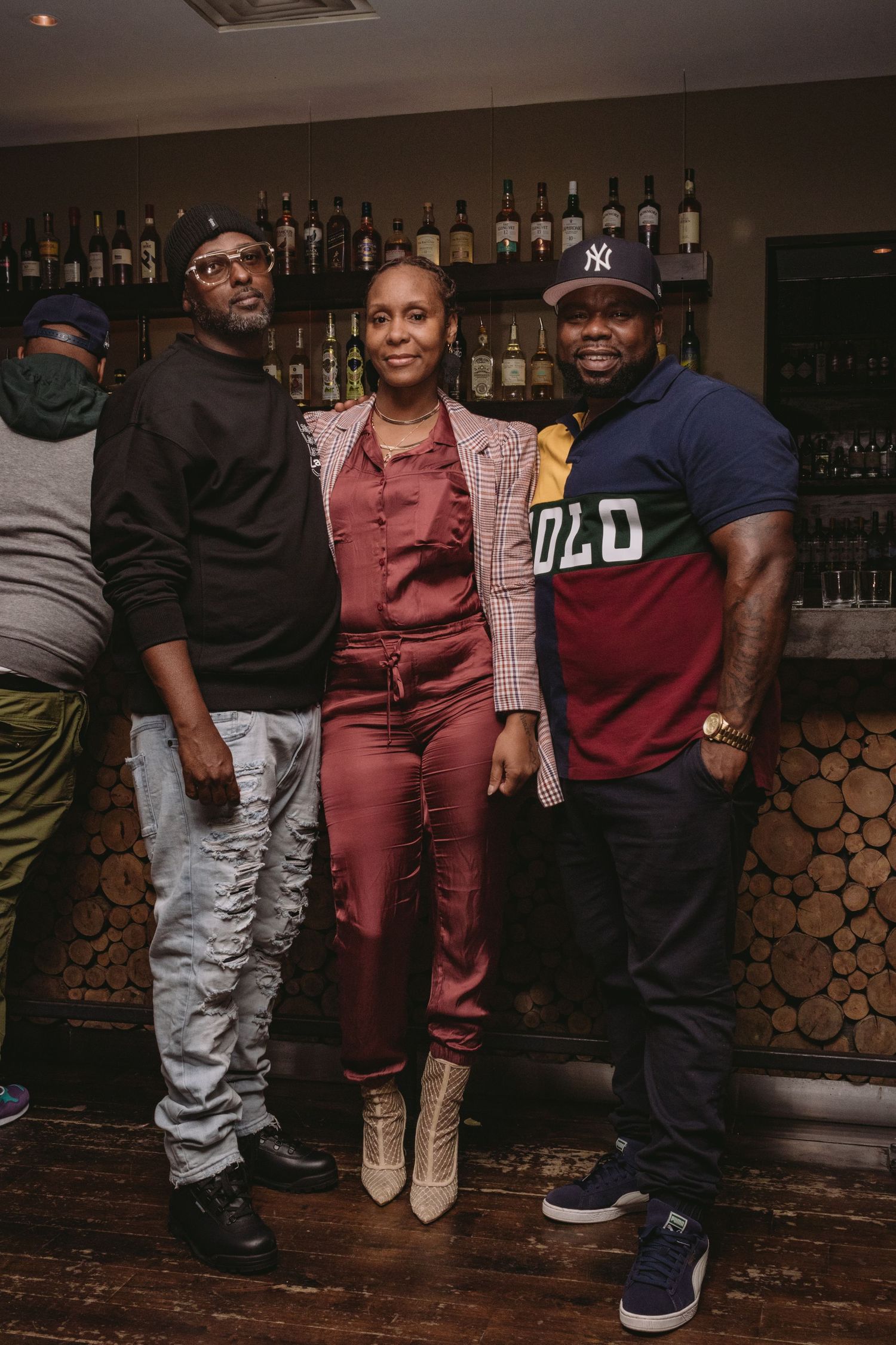 A group in casual evening attire poses together at a bar or lounge with exposed brick walls and wooden accents.