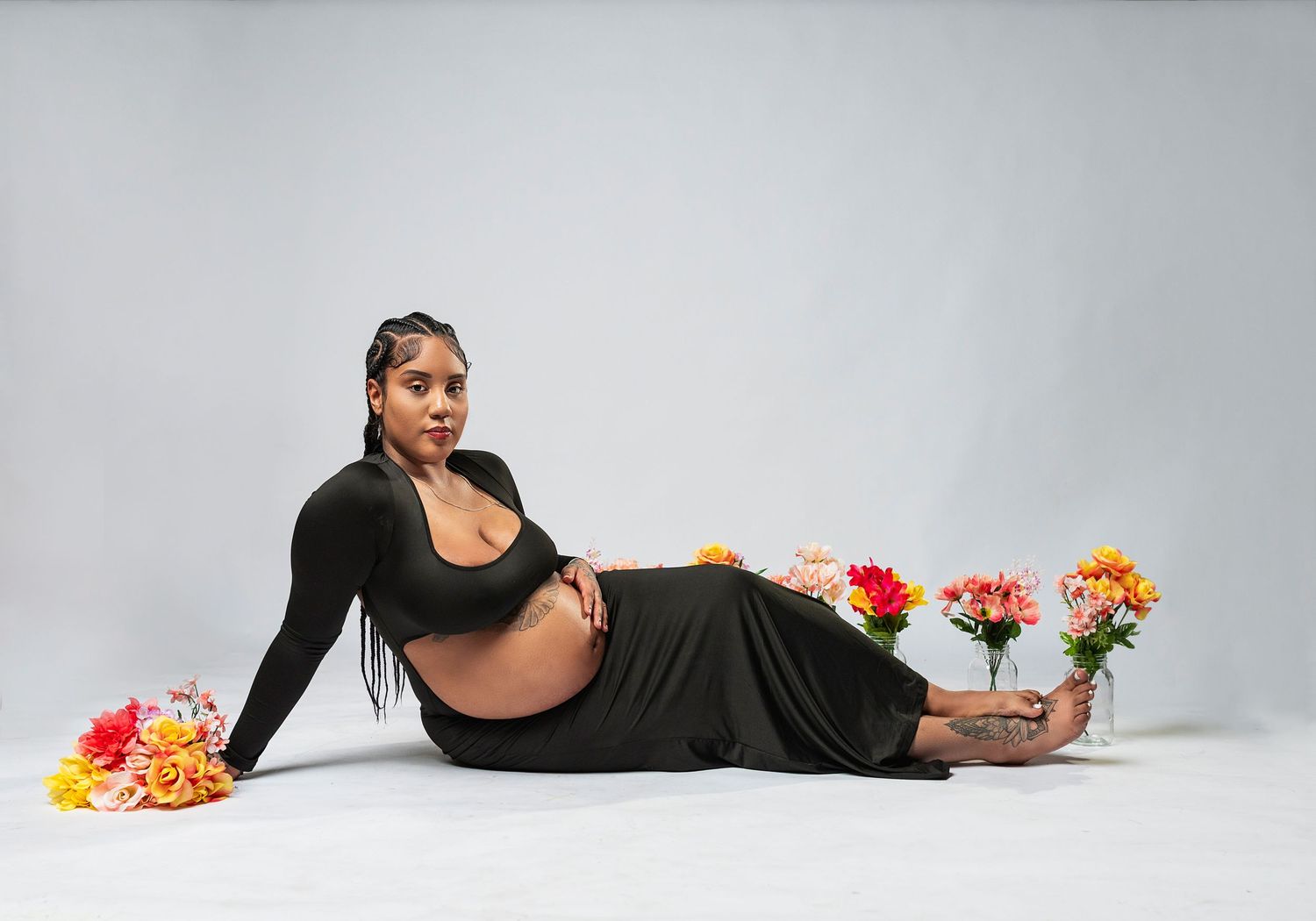 Maternity photo with woman in black outfit reclining among colorful flowers against light background.