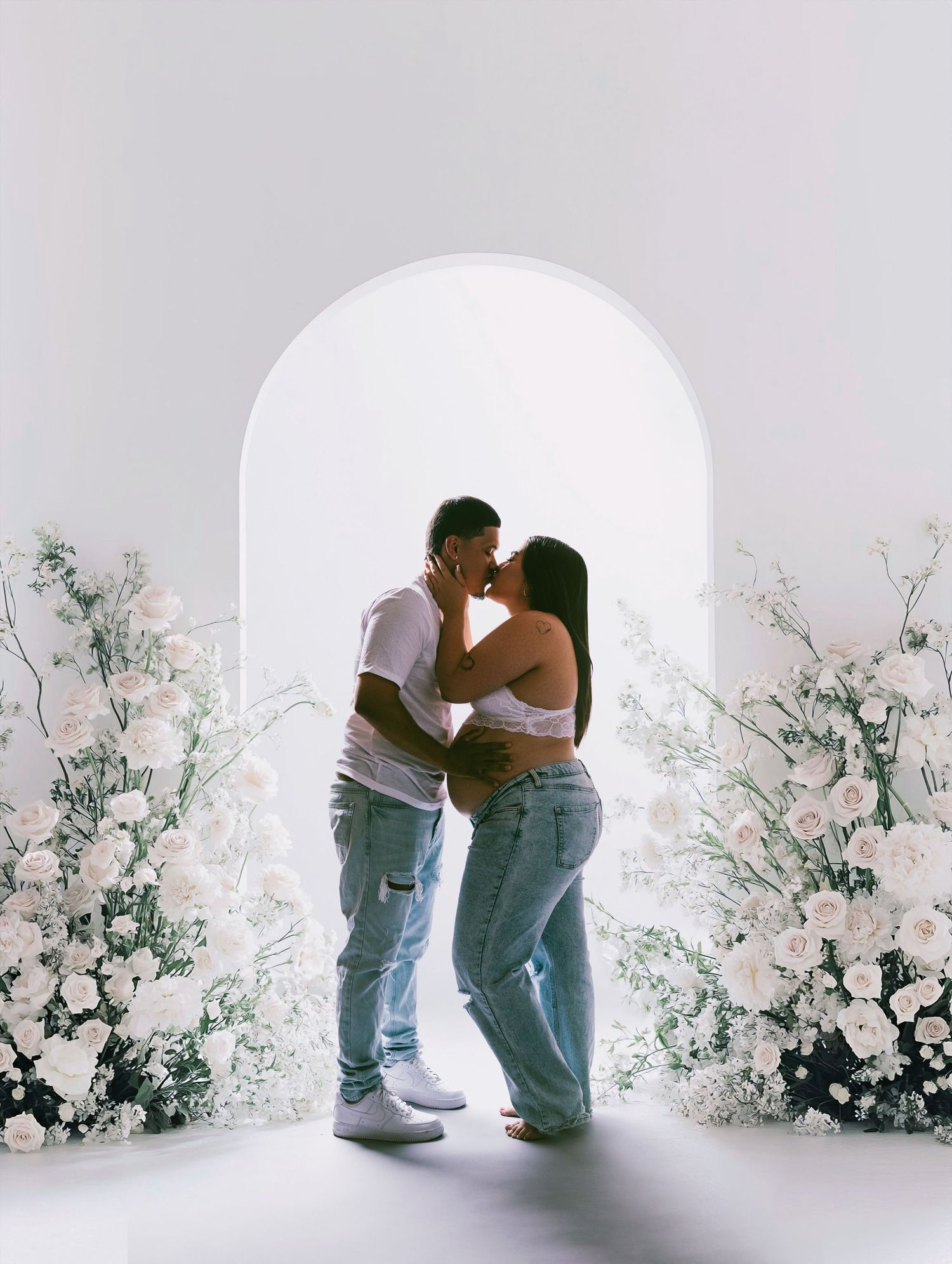 Couple kissing in an arch doorway surrounded by white floral arrangements in a bright, airy maternity photo shoot.