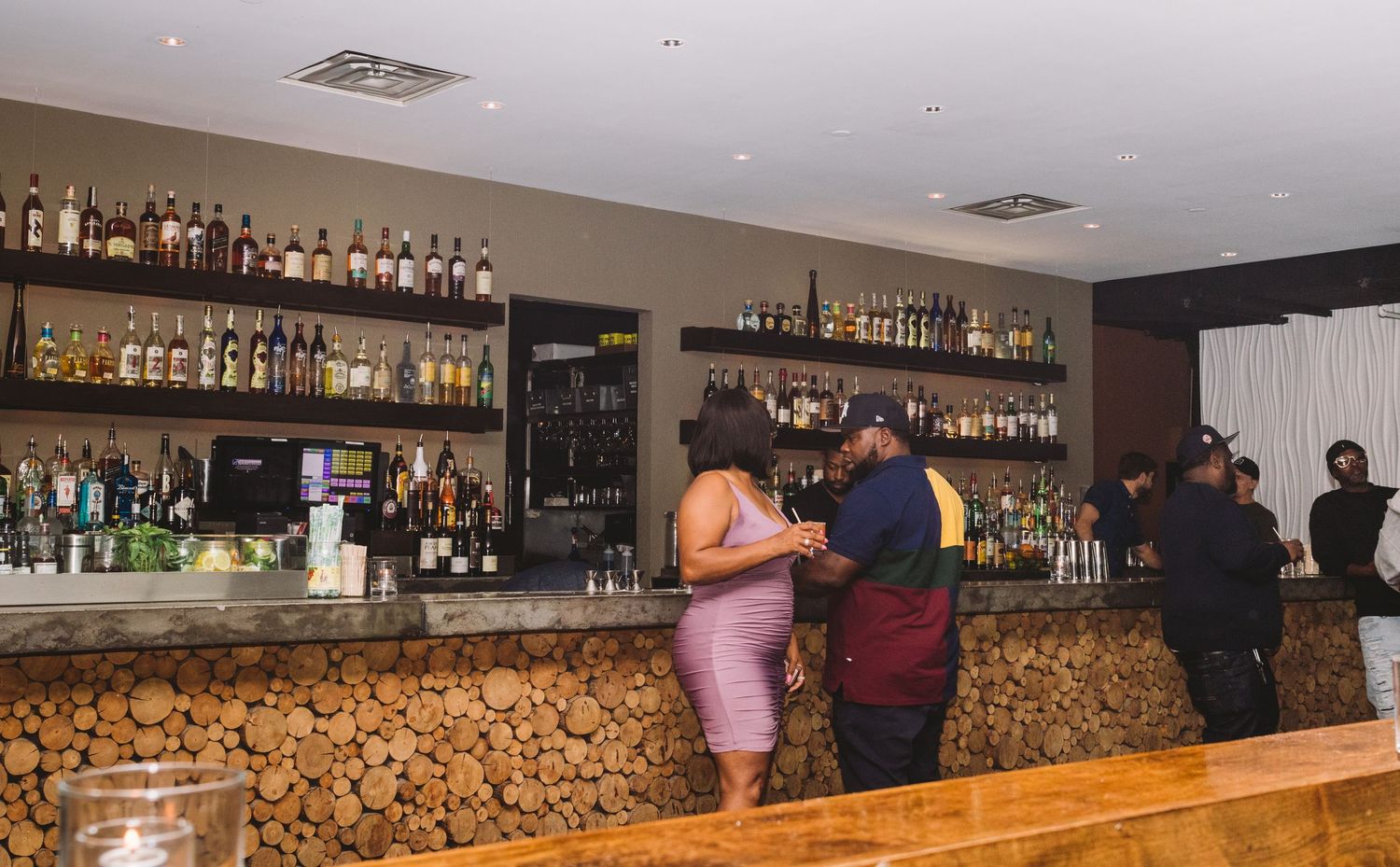 Modern bar interior with wooden counter decorated with cork panels and extensive liquor bottle display on shelves.