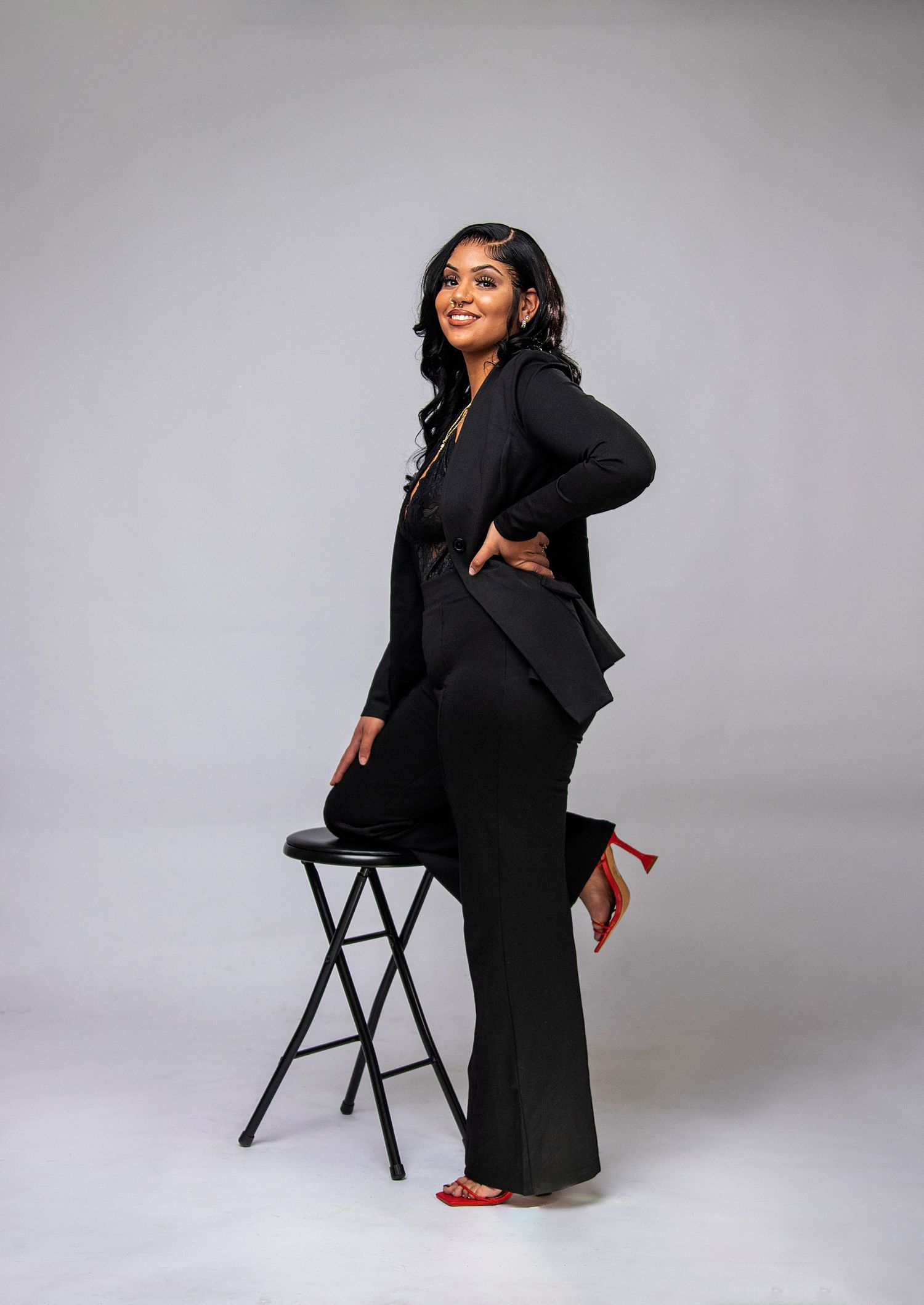 A professional portrait featuring a black pantsuit and red heels against a grey studio background.