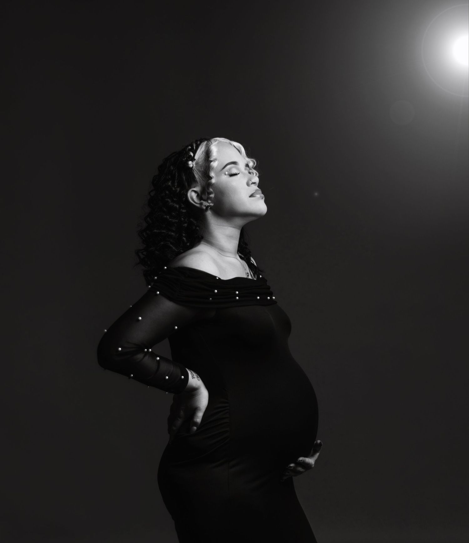 Dramatic black and white maternity portrait with woman in sparkly dress against dark background.