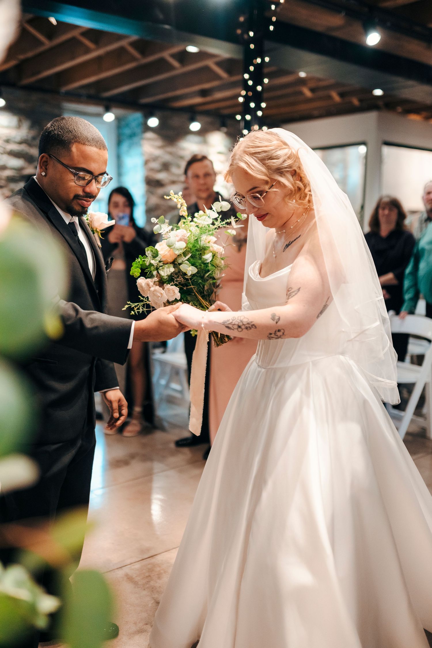 Bride in white gown with veil and groom in black suit exchange bouquet during indoor wedding ceremony.