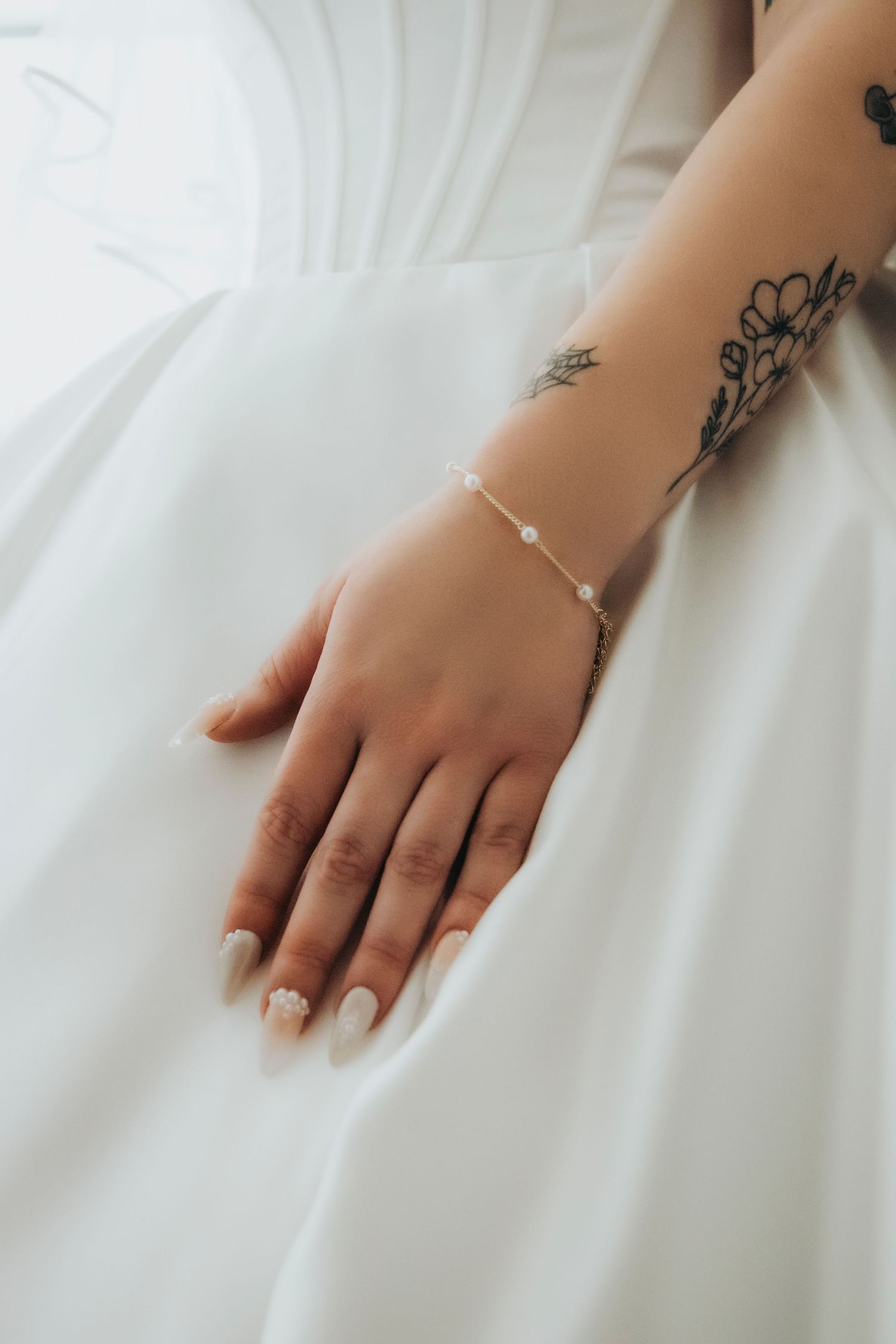 Bride's hand resting on white wedding dress showcasing delicate pearl bracelet and floral wrist tattoo.