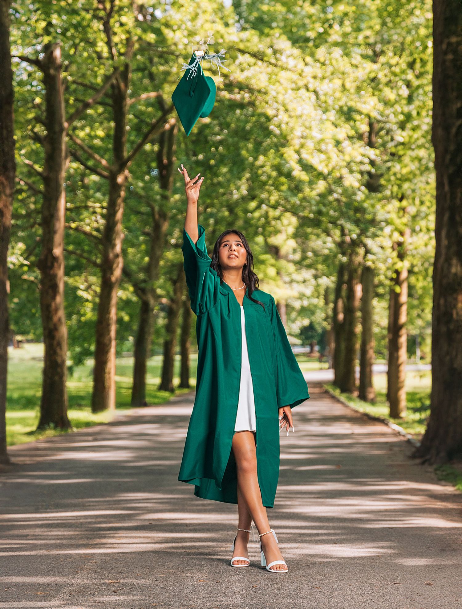Graduate in emerald green gown tosses cap on tree-lined path during celebration photo.